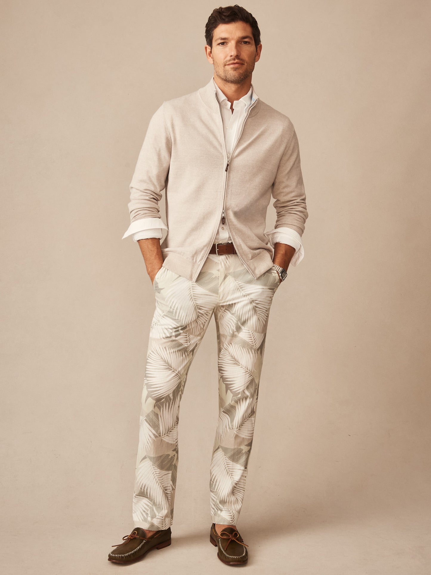 Man wearing an Orwell Sweater by J.McLaughlin in a neutral tone, paired with palm leaf patterned pants and loafers, showcasing effortlessly stylish layering with enduring designs.