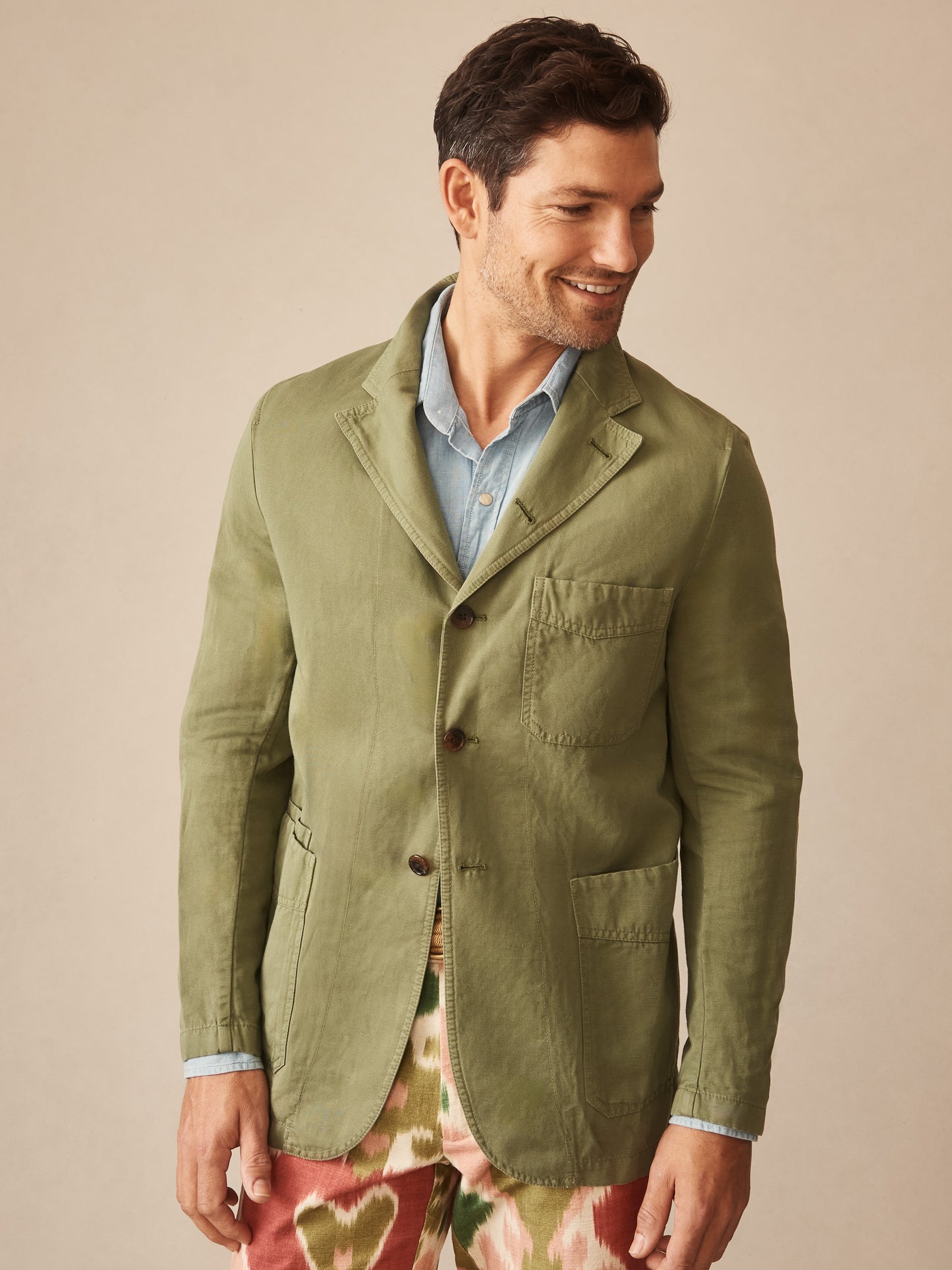 Man wearing a sage green Charlestown Jacket by J.McLaughlin over a light blue shirt, paired with patterned pants, showcasing effortlessly stylish and enduring designs for a life well-lived.