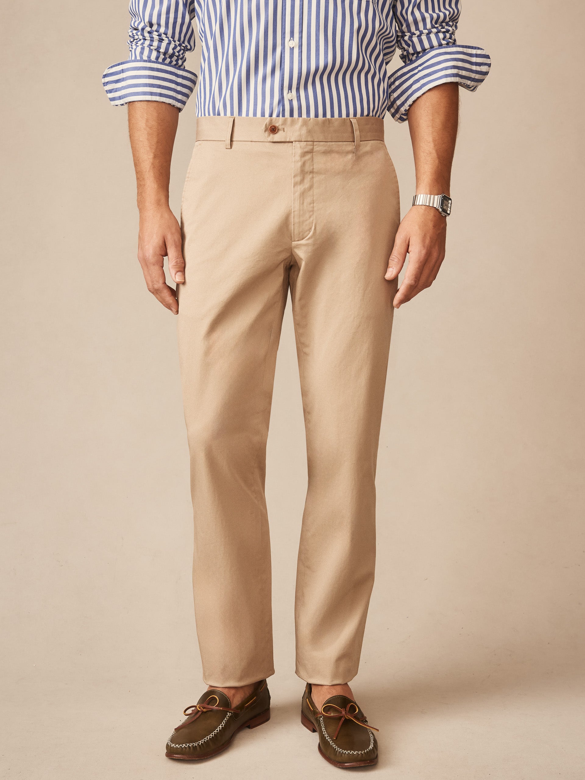 Man wearing beige Madison Pants by J.McLaughlin paired with a blue and white striped shirt and brown loafers, showcasing effortlessly stylish, enduring designs.