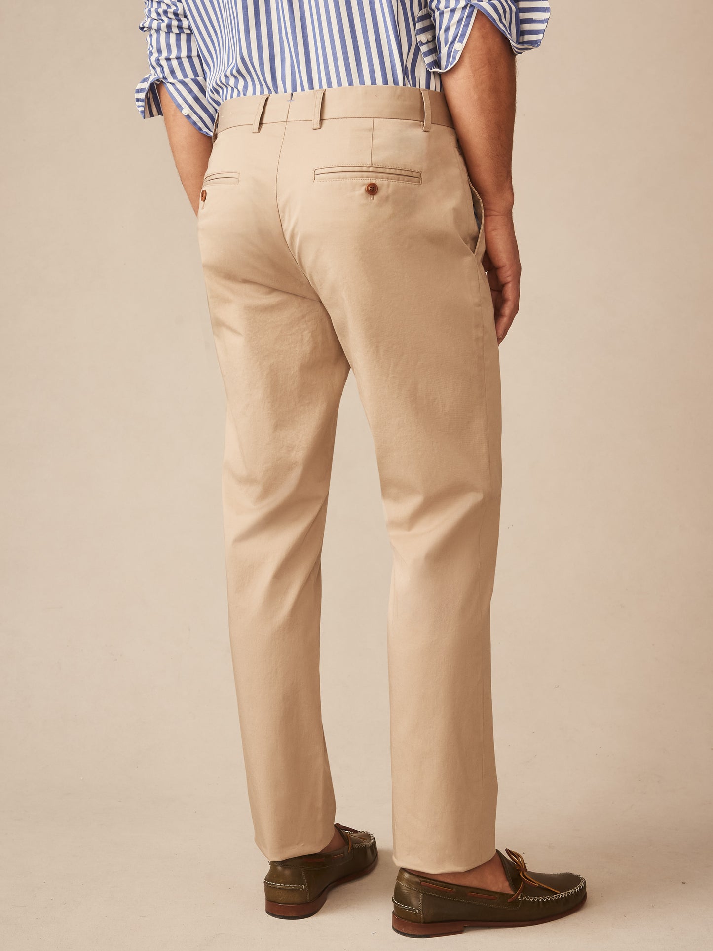 Back view of a man wearing beige Madison Pants by J.McLaughlin paired with loafers and a blue striped shirt cuffed at the sleeves, showcasing effortlessly stylish, enduring designs in innovative fabrics for a legacy of timeless fashion. A destination for defining style. Life, Well-Lived.