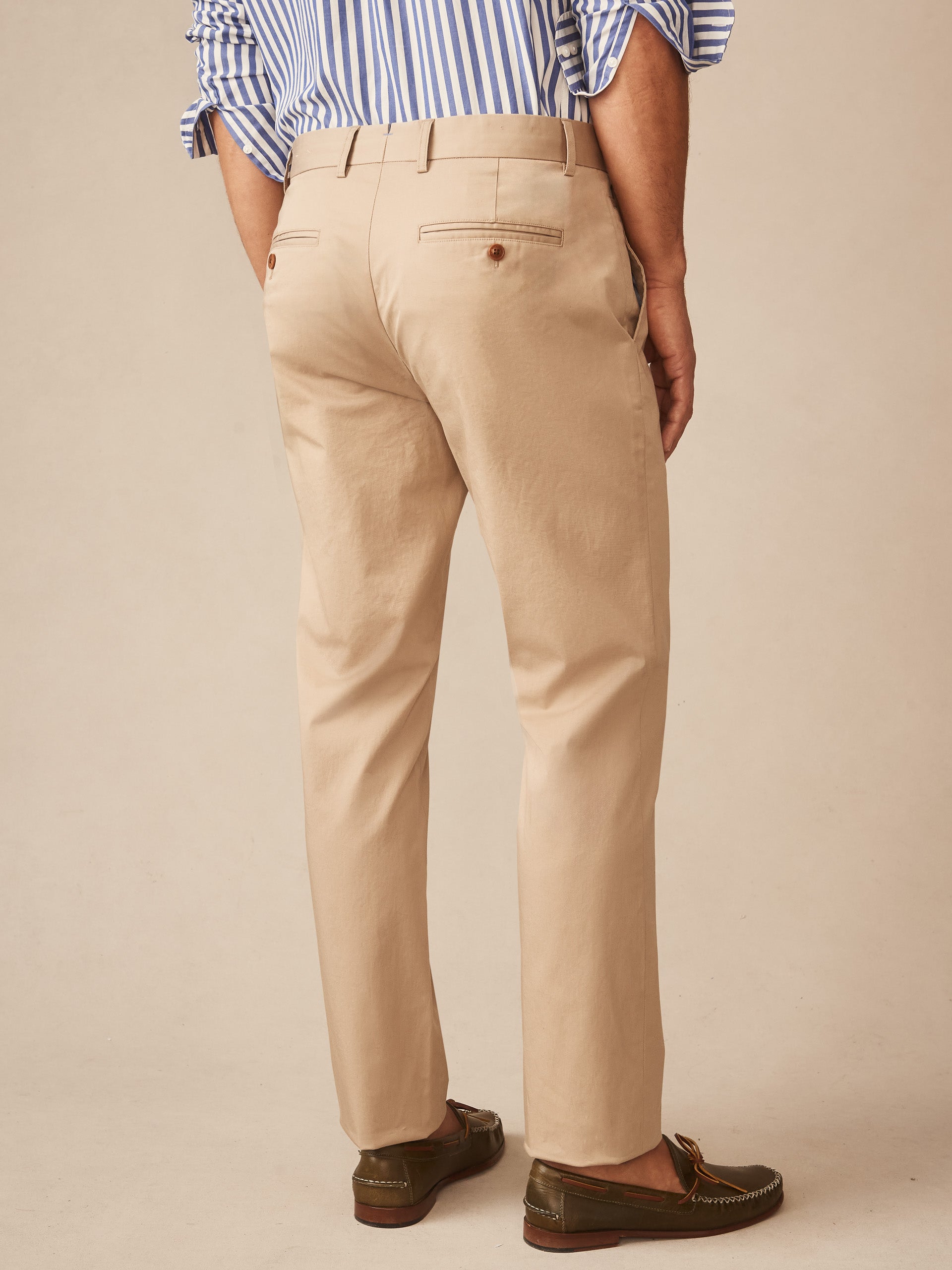 Back view of a man wearing beige Madison Pants by J.McLaughlin paired with loafers and a blue striped shirt cuffed at the sleeves, showcasing effortlessly stylish, enduring designs in innovative fabrics for a legacy of timeless fashion. A destination for defining style. Life, Well-Lived.