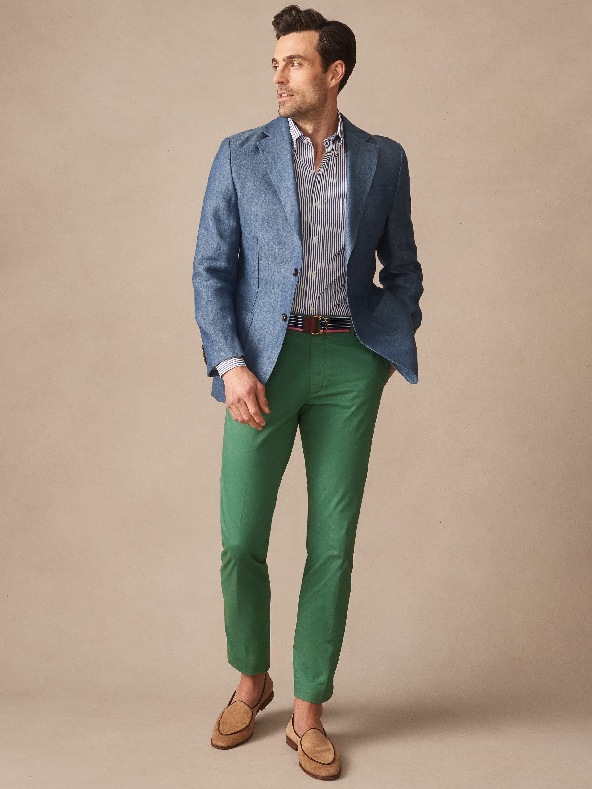 Man wearing a Brera Linen Sport Coat in Herringbone by J.McLaughlin paired with green pants, a striped shirt, and loafers, showcasing effortlessly stylish, enduring designs and innovative fabrics.