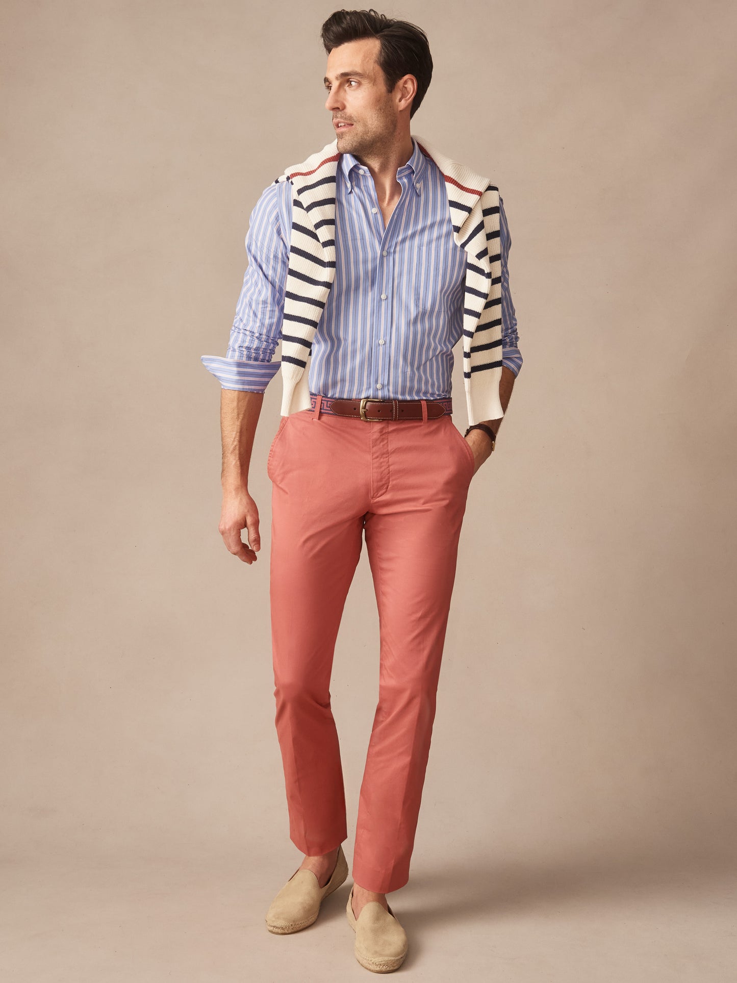 Man wearing J.McLaughlin Madison Pants in a warm coral tone, paired with a blue striped shirt and a casual striped sweater draped over shoulders, showcasing effortlessly stylish, enduring designs.