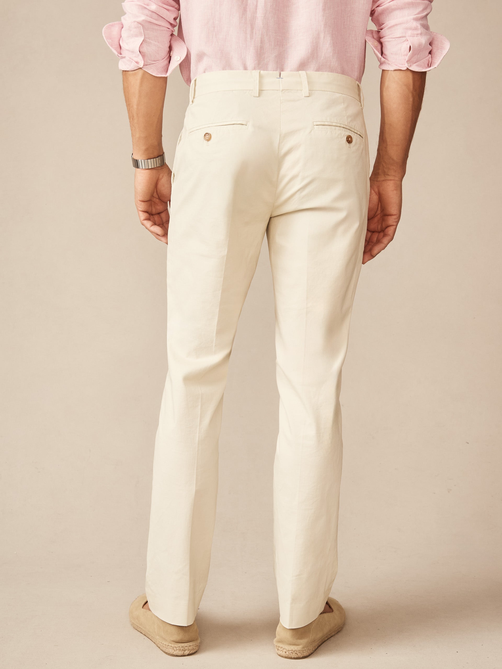 Back view of a man wearing stone-colored Madison Pants by J.McLaughlin paired with a rolled-up pink shirt and neutral shoes, showcasing effortlessly stylish and enduring designs in innovative fabrics.