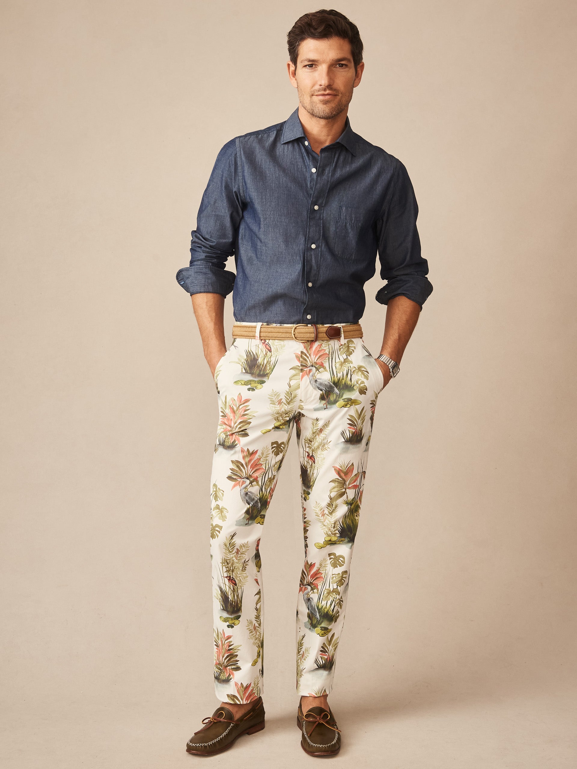 Man wearing a navy blue button-up shirt paired with J.McLaughlin Madison Fancy Pants in Tidal Palms featuring a tropical print, styled with a tan belt and brown loafers, showcasing effortlessly stylish, enduring designs and innovative fabrics from a legacy of timeless fashion. A destination for defining style. Life, Well-Lived.