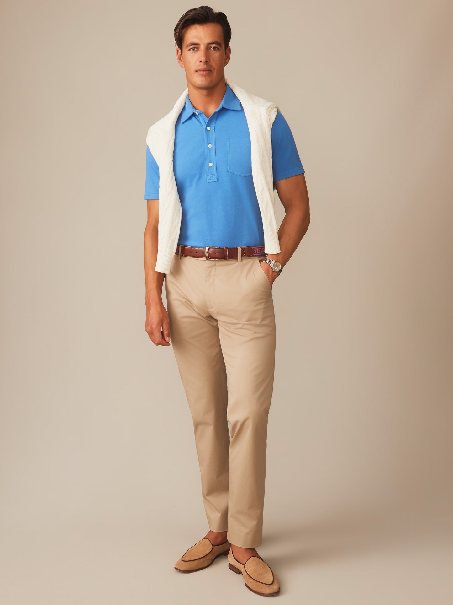 Male model wearing a Pacific blue Mantle Pique Polo by J.McLaughlin paired with beige pants, a white sweater draped over shoulders, and tan loafers for an effortlessly stylish look.