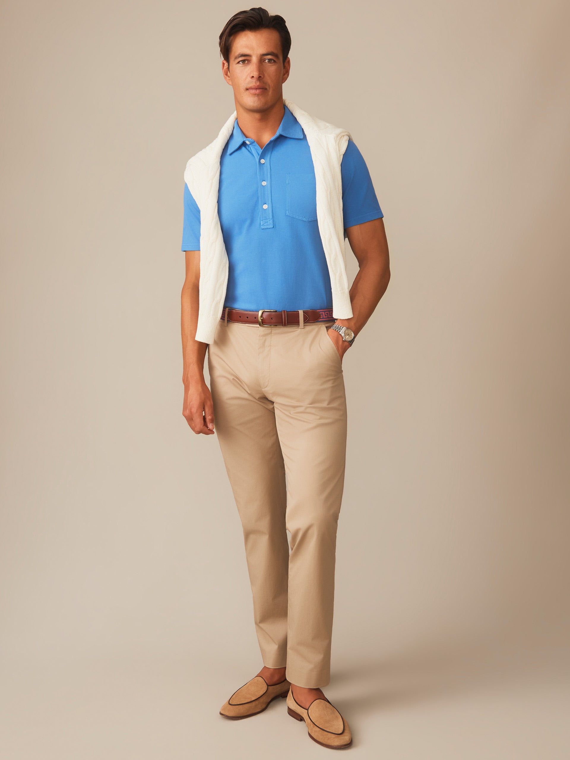 Male model wearing a Pacific blue Mantle Pique Polo by J.McLaughlin paired with beige pants, a white sweater draped over shoulders, and tan loafers for an effortlessly stylish look.