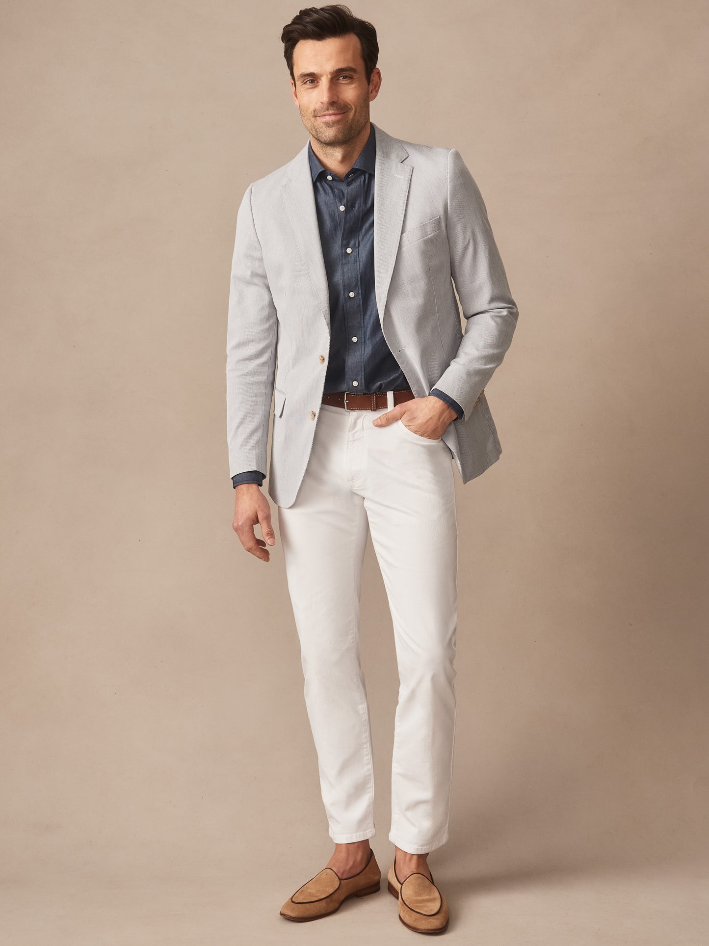 Man wearing a light gray Suffolk Sport Coat in Fineline by J.McLaughlin over a dark blue shirt with white pants and tan loafers, standing against a neutral background.