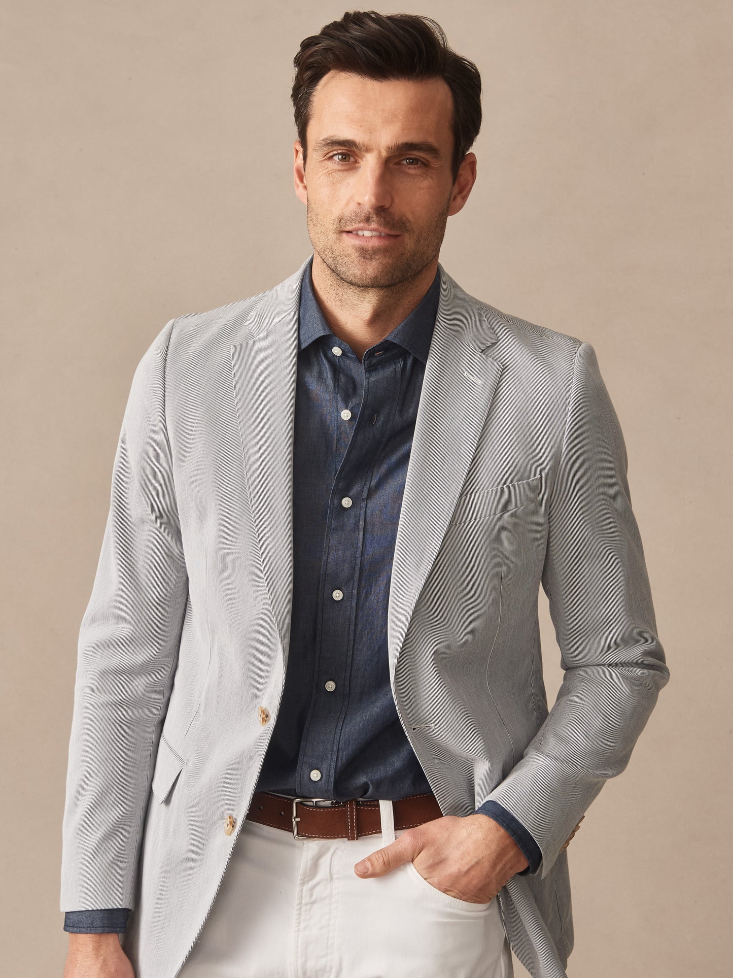 Man wearing a light gray Suffolk Sport Coat in Fineline over a dark blue shirt with white pants and brown belt, showcasing J.McLaughlin’s timeless tailored jacket.