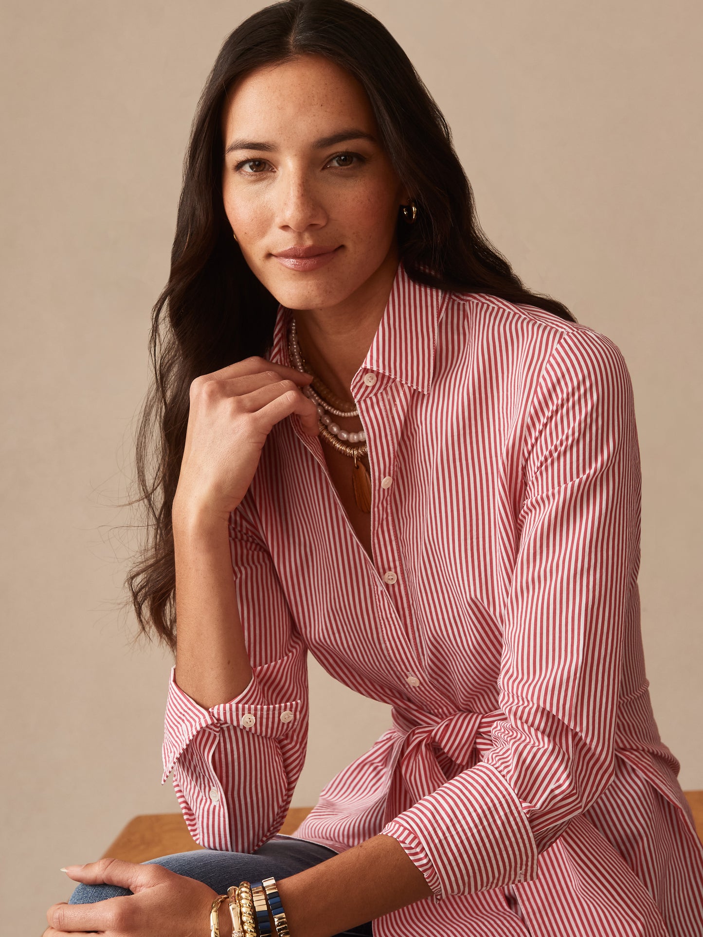 Woman wearing a Margaux Shirt in Stripe by J.McLaughlin with red and white vertical stripes, styled with layered necklaces and bracelets, showcasing effortlessly stylish, enduring designs and innovative fabrics in a legacy of timeless fashion. A destination for defining style. Life, Well-Lived.