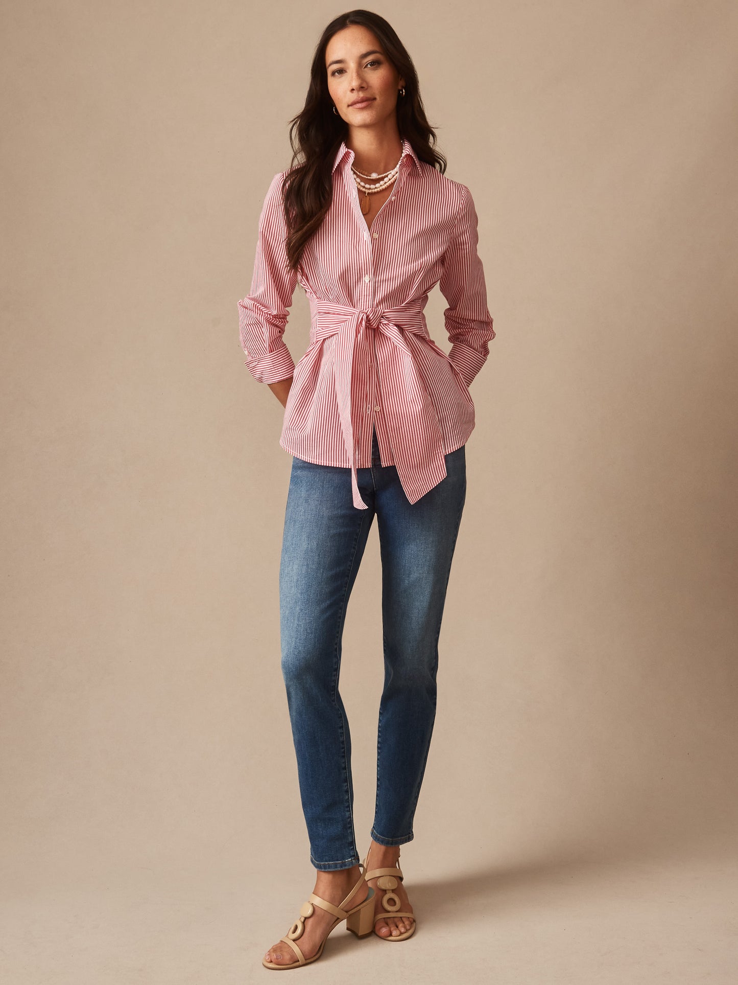 A woman wearing the Margaux Shirt in Stripe by J.McLaughlin in soft coral and white stripes, paired with jeans and sandals, showcasing effortlessly stylish, enduring designs and a legacy of timeless fashion. A destination for defining style. Life, Well-Lived.