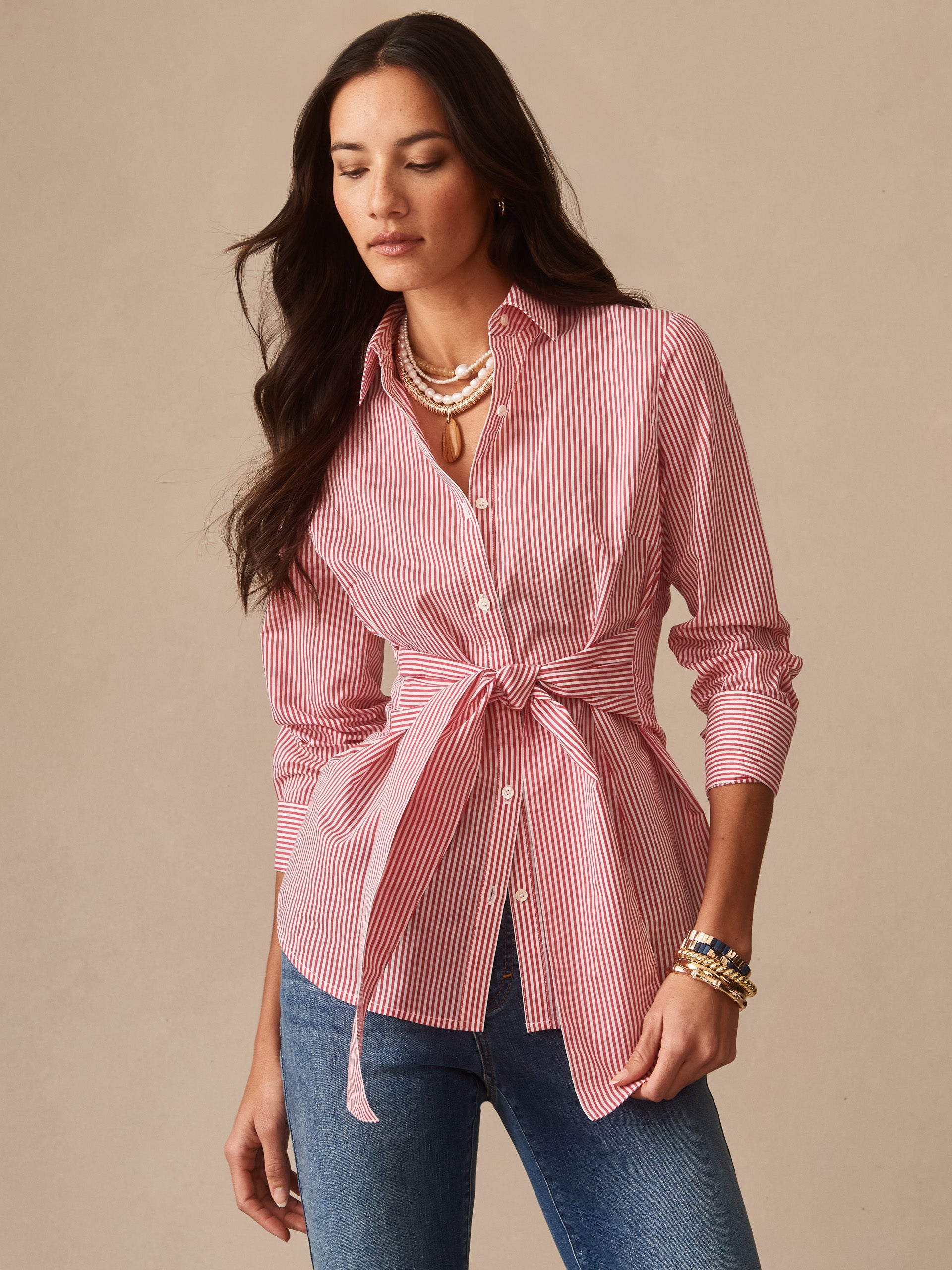 Woman wearing a Margaux Shirt in Stripe by J.McLaughlin, featuring red and white stripes with a tie waist, paired with layered necklaces and bracelets, embodying effortlessly stylish, enduring designs.