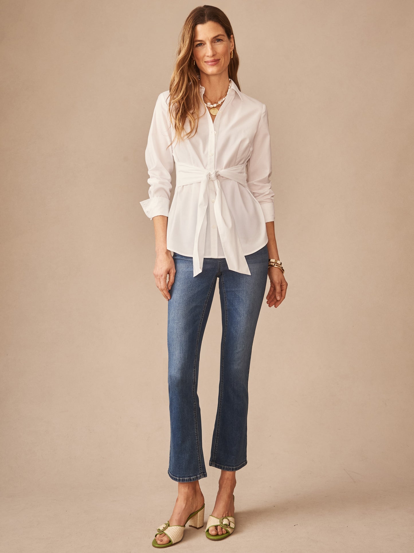 Woman models a white Margaux Shirt by J.McLaughlin, featuring a tied waist and classic collar, paired with blue jeans and stylish sandals, embodying effortlessly stylish, enduring designs.
