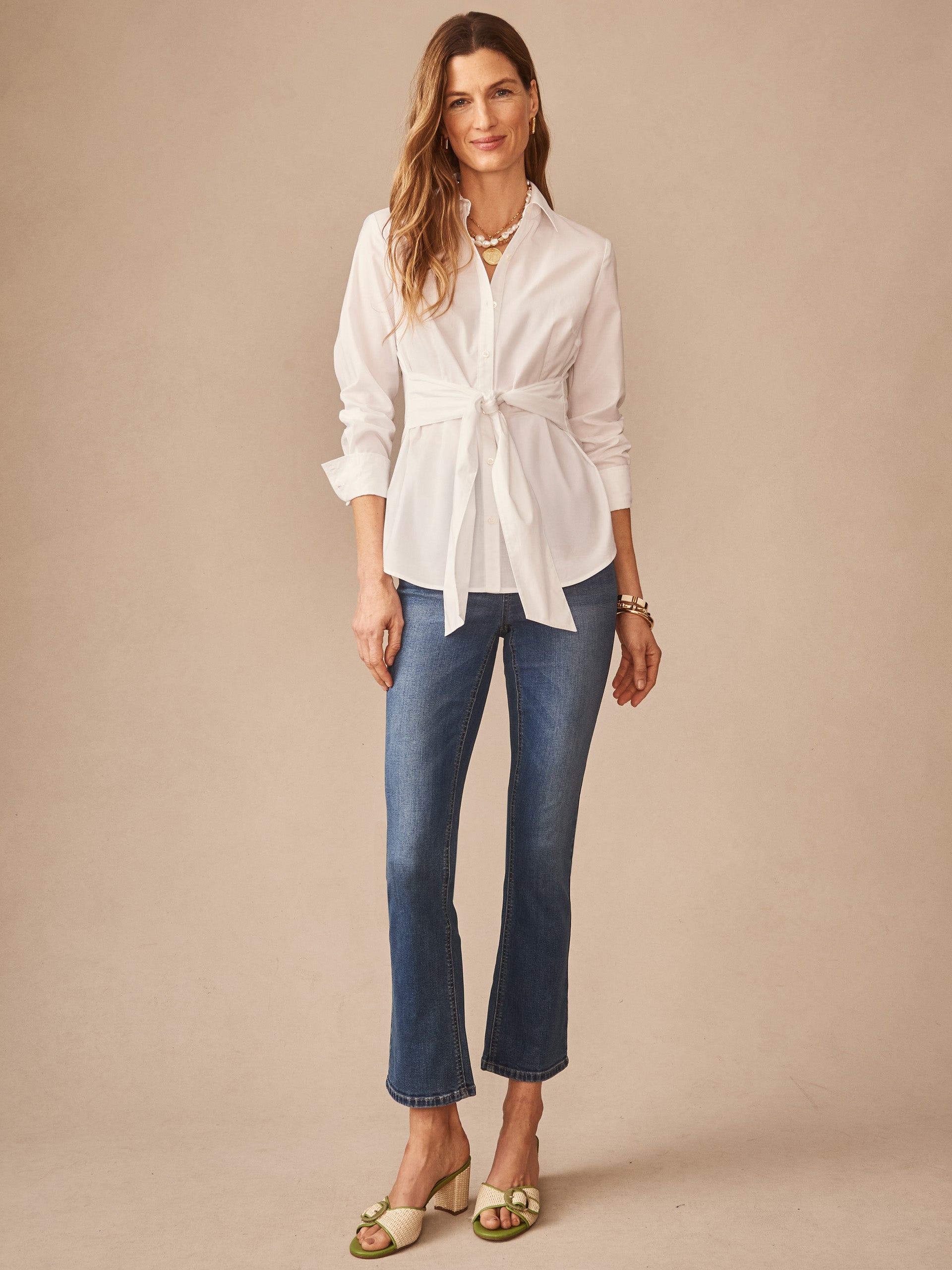 Woman models a white Margaux Shirt by J.McLaughlin, featuring a tied waist and classic collar, paired with blue jeans and stylish sandals, embodying effortlessly stylish, enduring designs.