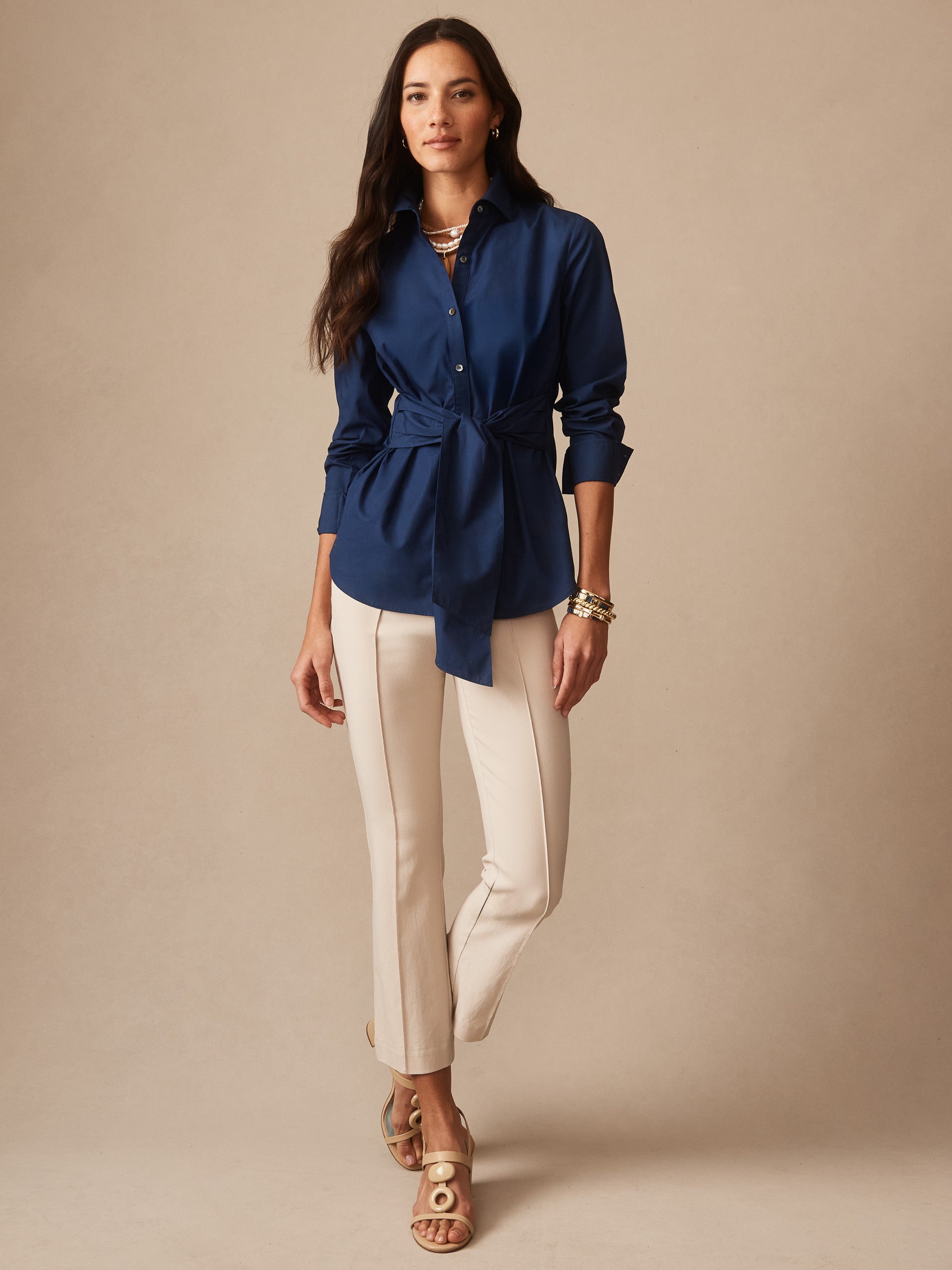 Woman wearing a navy Margaux Shirt by J.McLaughlin featuring a tie waist detail, paired with cream-colored fitted pants and beige sandals, showcasing effortlessly stylish, enduring designs.