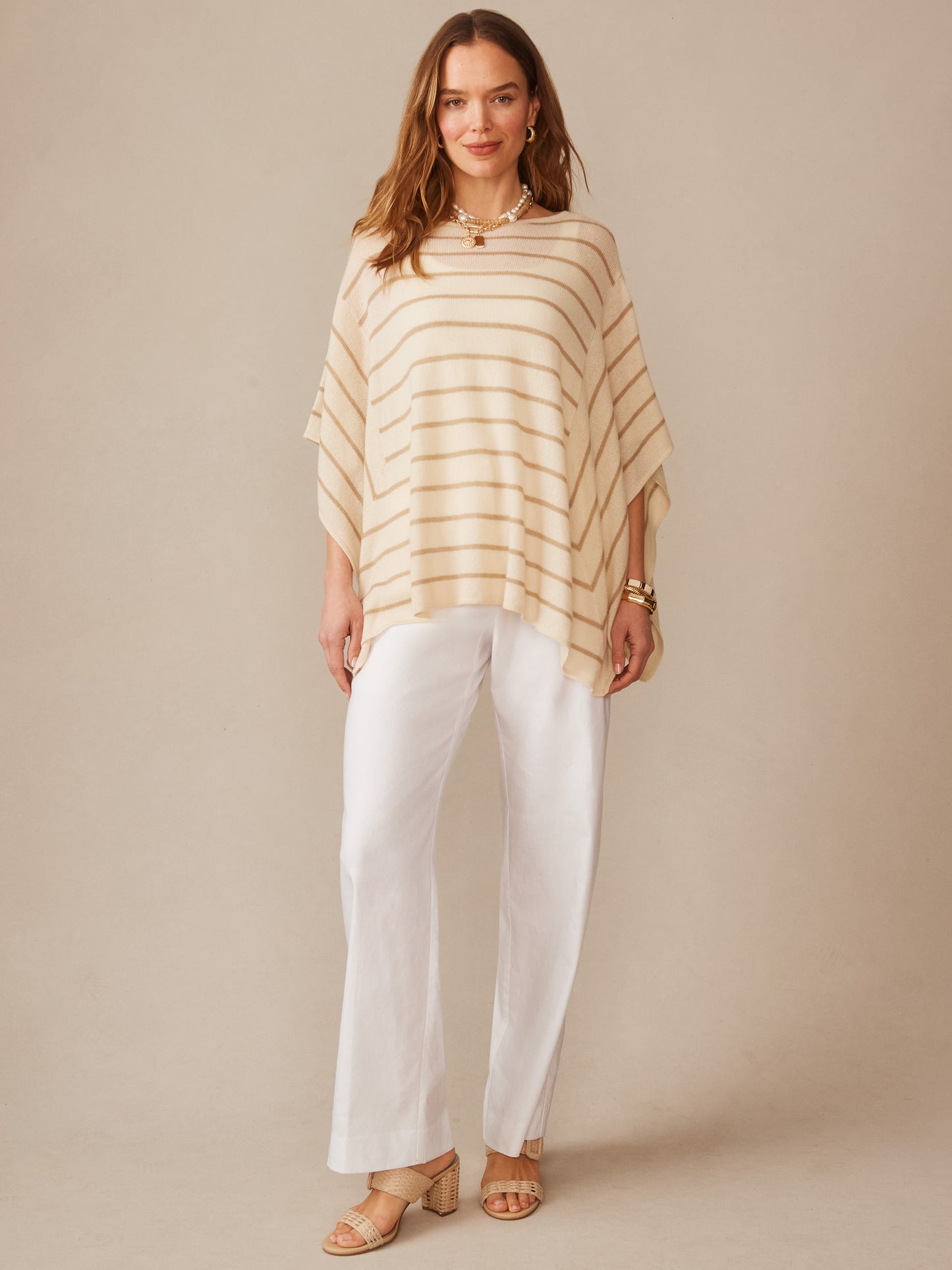 Woman wearing a striped Marin Cashmere Poncho in Stripe by J.McLaughlin, paired with white pants and sandals, showcasing effortlessly stylish and enduring designs.