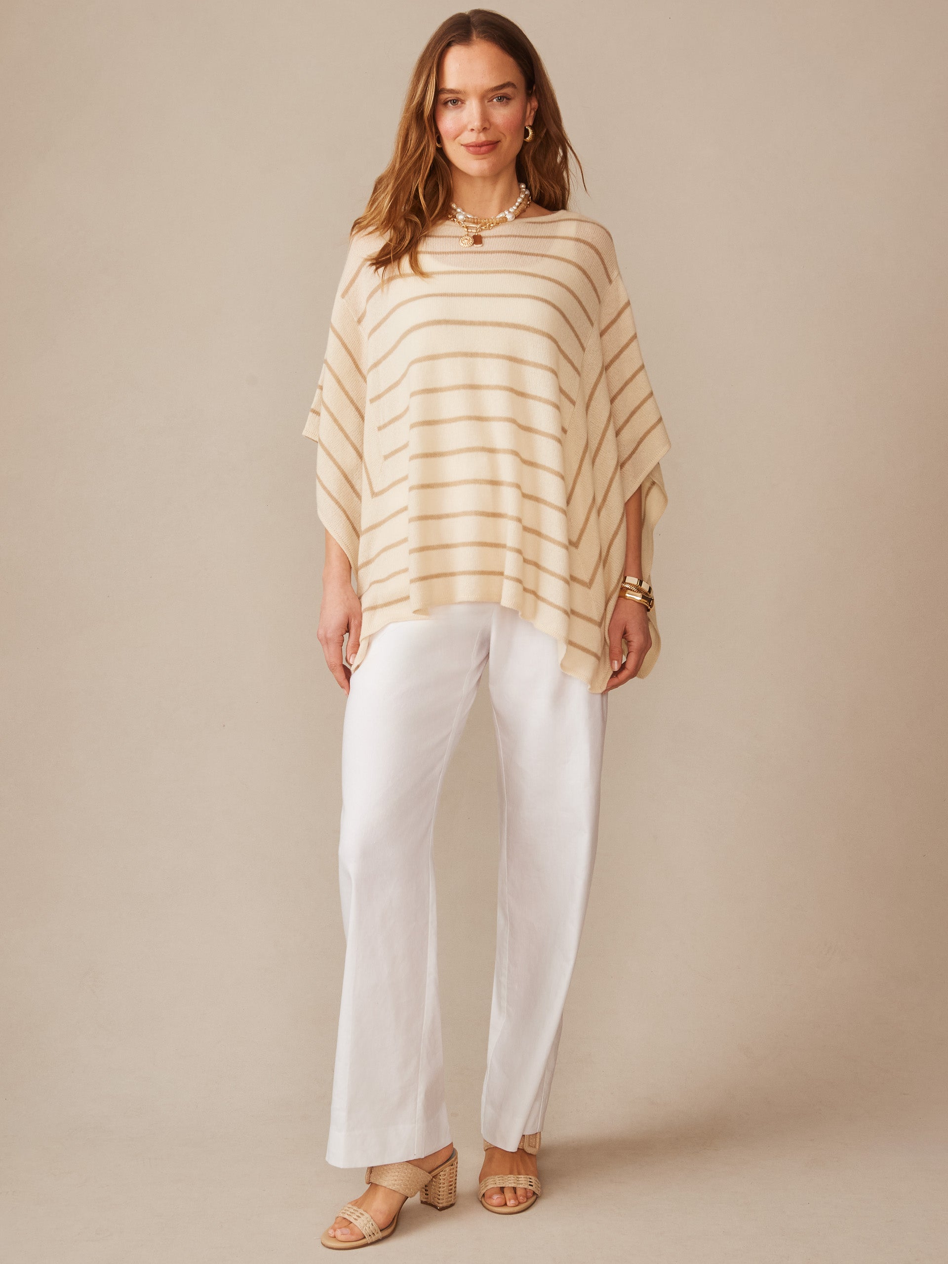 Woman wearing a striped Marin Cashmere Poncho in Stripe by J.McLaughlin, paired with white pants and sandals, showcasing effortlessly stylish and enduring designs.