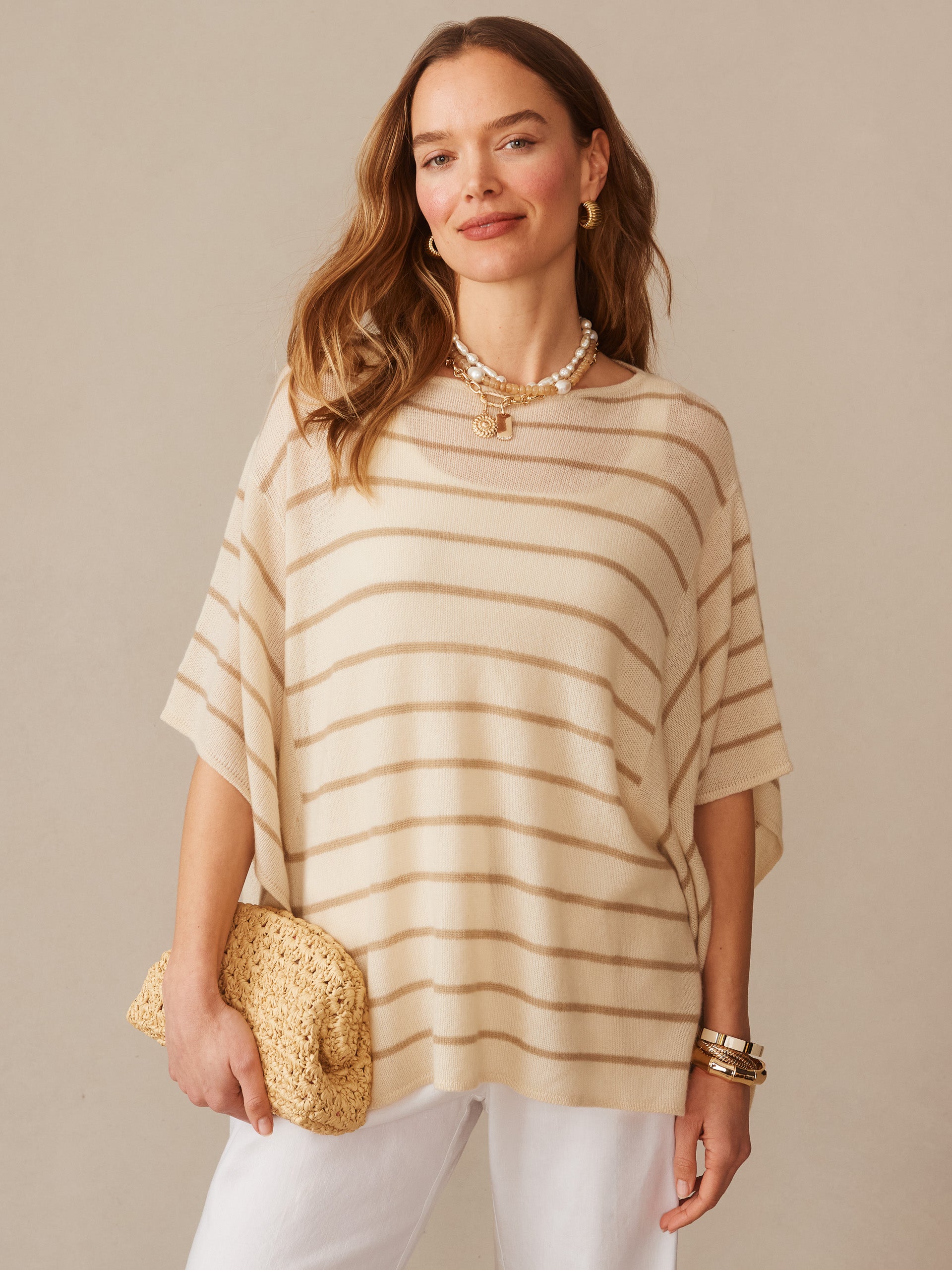 Woman wearing a Marin Cashmere Poncho in Stripe by J.McLaughlin featuring soft beige and sand dollar stripes, styled with white pants and gold accessories for an effortlessly stylish look.