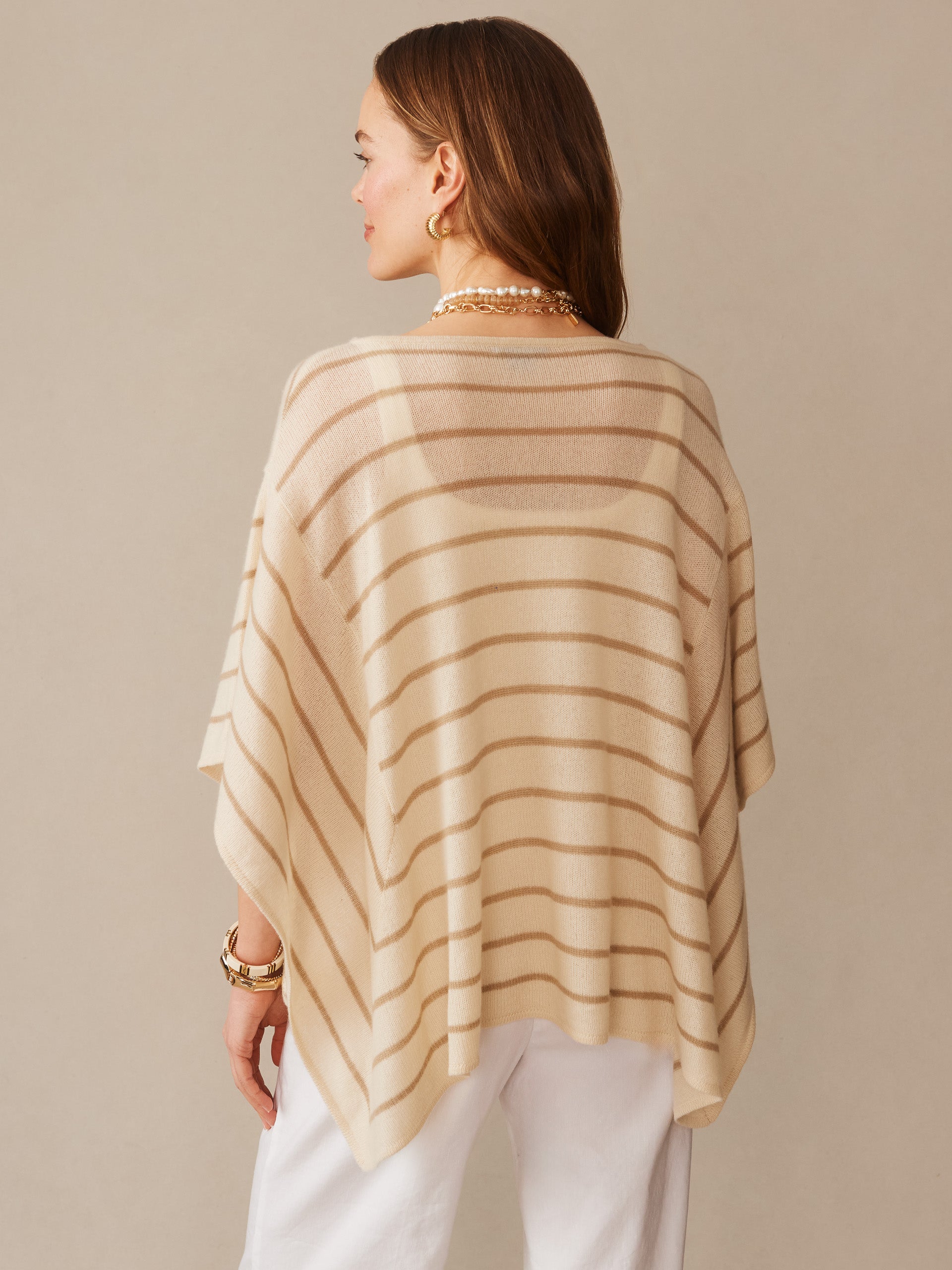Woman wearing a J.McLaughlin Marin Cashmere Poncho in Stripe, featuring soft beige and sand dollar horizontal stripes, paired with white pants and gold accessories, showcasing effortlessly stylish, enduring designs and innovative fabrics in a timeless fashion look.