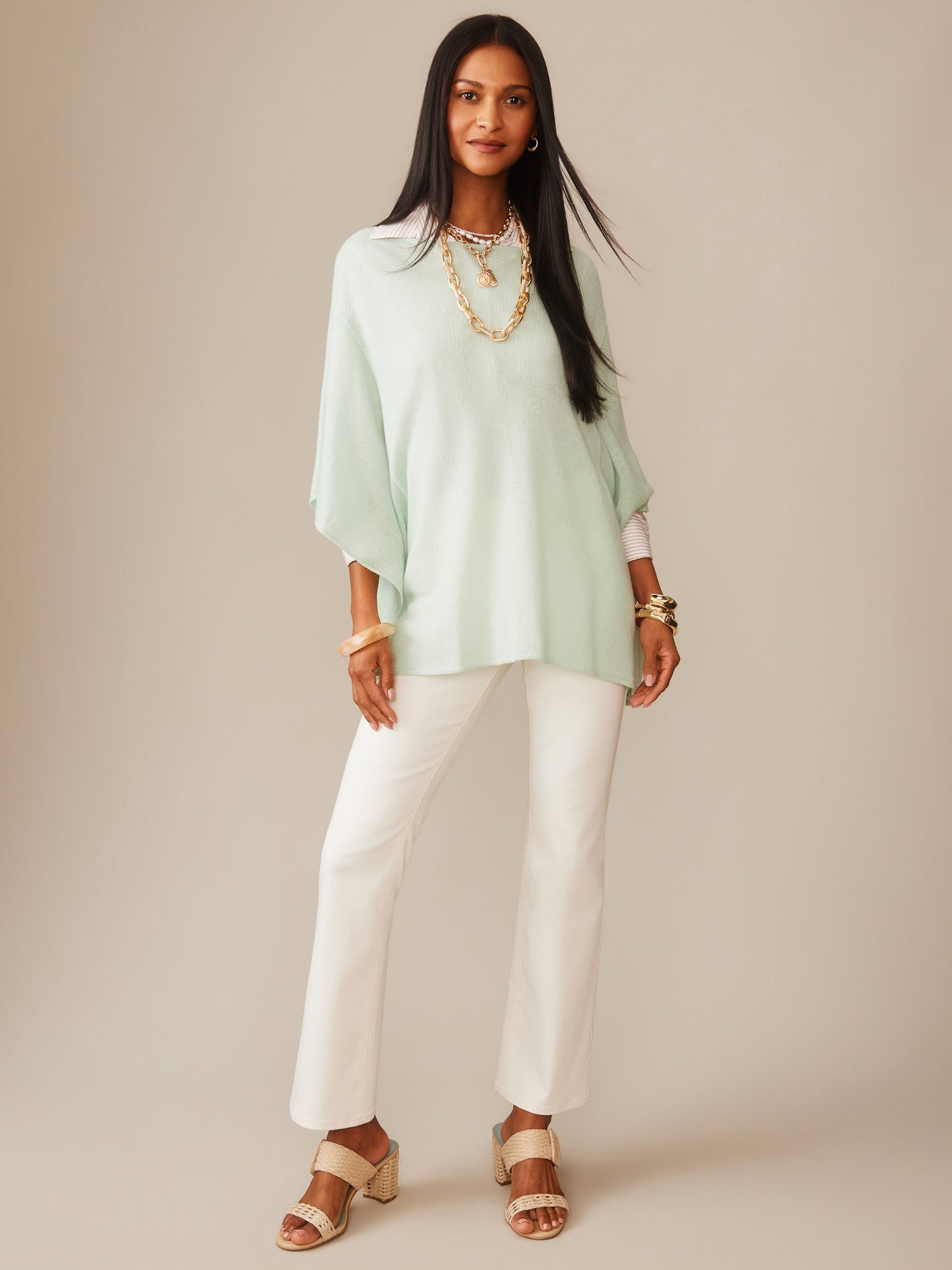 Woman wearing a light mint Marin Cashmere Poncho by J.McLaughlin paired with white pants and beige sandals, showcasing effortlessly stylish, enduring designs and a legacy of timeless fashion.