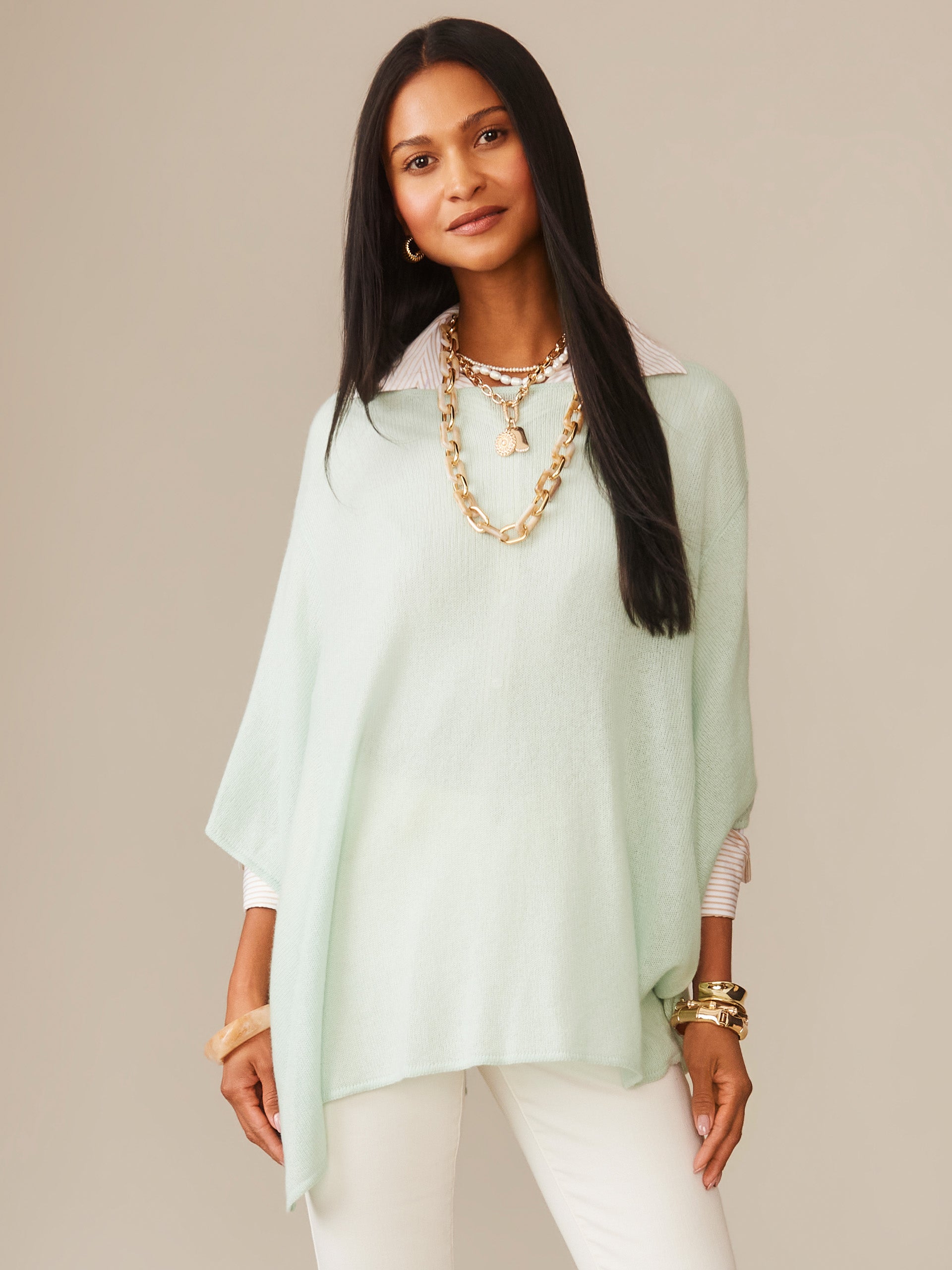 A woman wearing a light mint Marin Cashmere Poncho by J.McLaughlin, styled with layered gold necklaces, bracelets, and white pants, embodying effortlessly stylish, enduring designs in innovative fabrics.