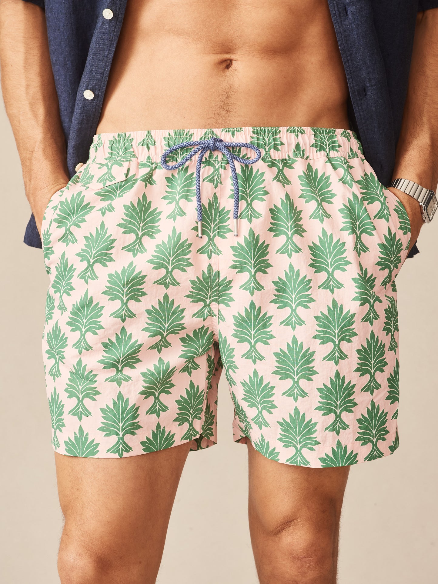 Close-up of a man wearing Marina Swim Trunks in Shadow Fronds by J.McLaughlin, featuring green leaf patterns on a light pink background, showcasing effortlessly stylish swimwear with enduring designs and innovative fabrics, reflecting a legacy of timeless fashion from a destination for defining style. Life, Well-Lived.