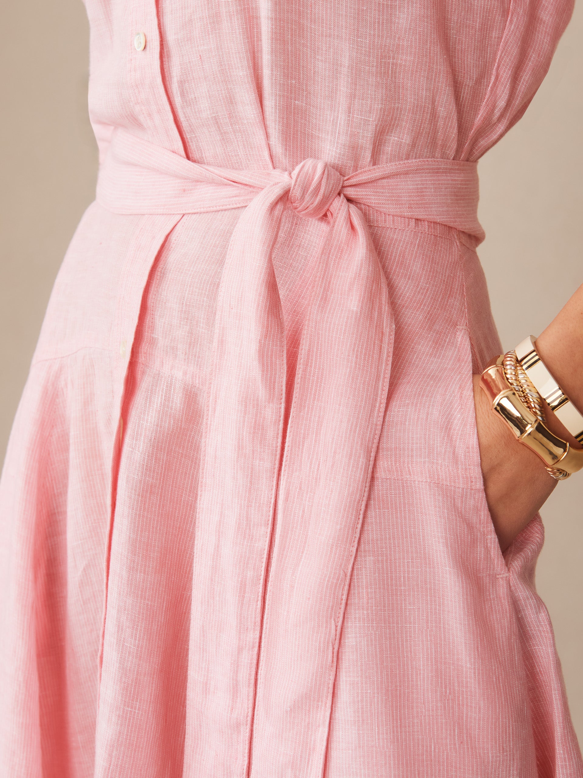 Close-up of a pink striped Mary Linen Dress in Stripe by J.McLaughlin featuring a tied waist belt and a hand resting in the dress pocket, showcasing effortlessly stylish, enduring designs with innovative fabrics.
