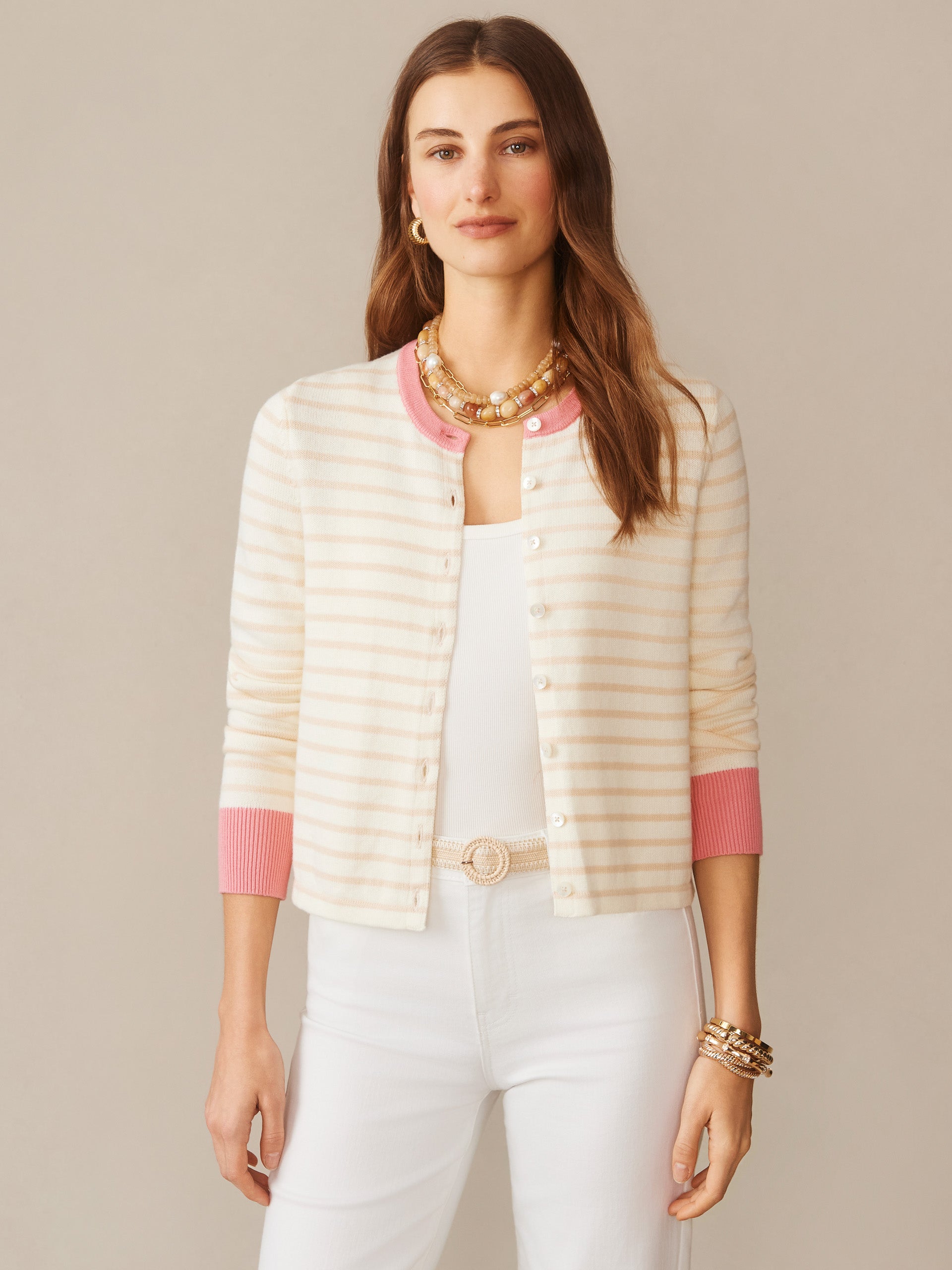Woman wearing a Matilda Cardigan in Stripe by J.McLaughlin, featuring soft off-white and oatmeal stripes with pink cuffs and collar, paired with white pants and gold accessories, showcasing effortlessly stylish, enduring designs and innovative fabrics in a legacy of timeless fashion. A destination for defining style. Life, Well-Lived.