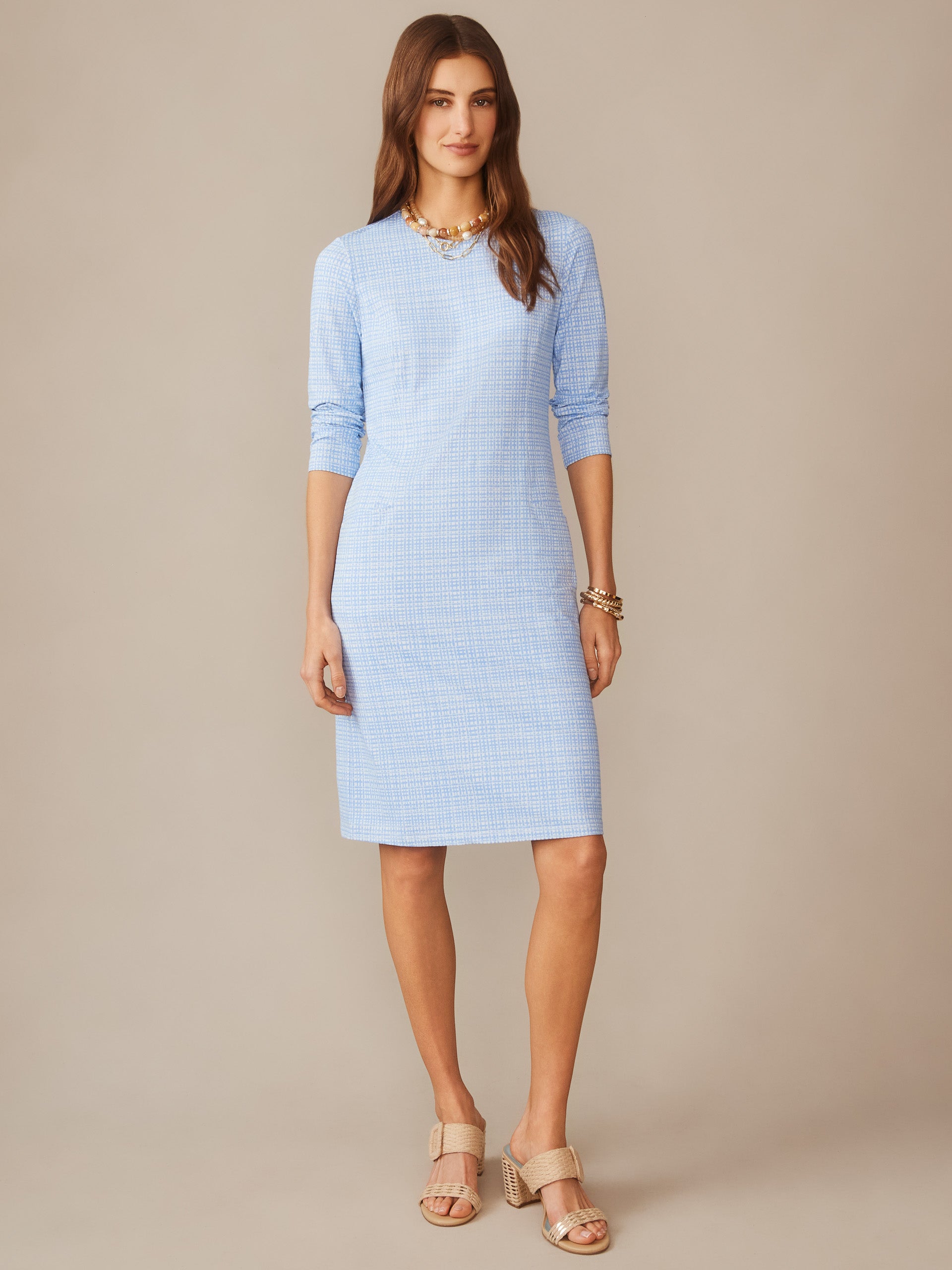 Woman wearing a light blue, subtly patterned Catalyst Dress in Pocket Square by J.McLaughlin, styled with layered gold necklace, bracelets, and beige heeled sandals.