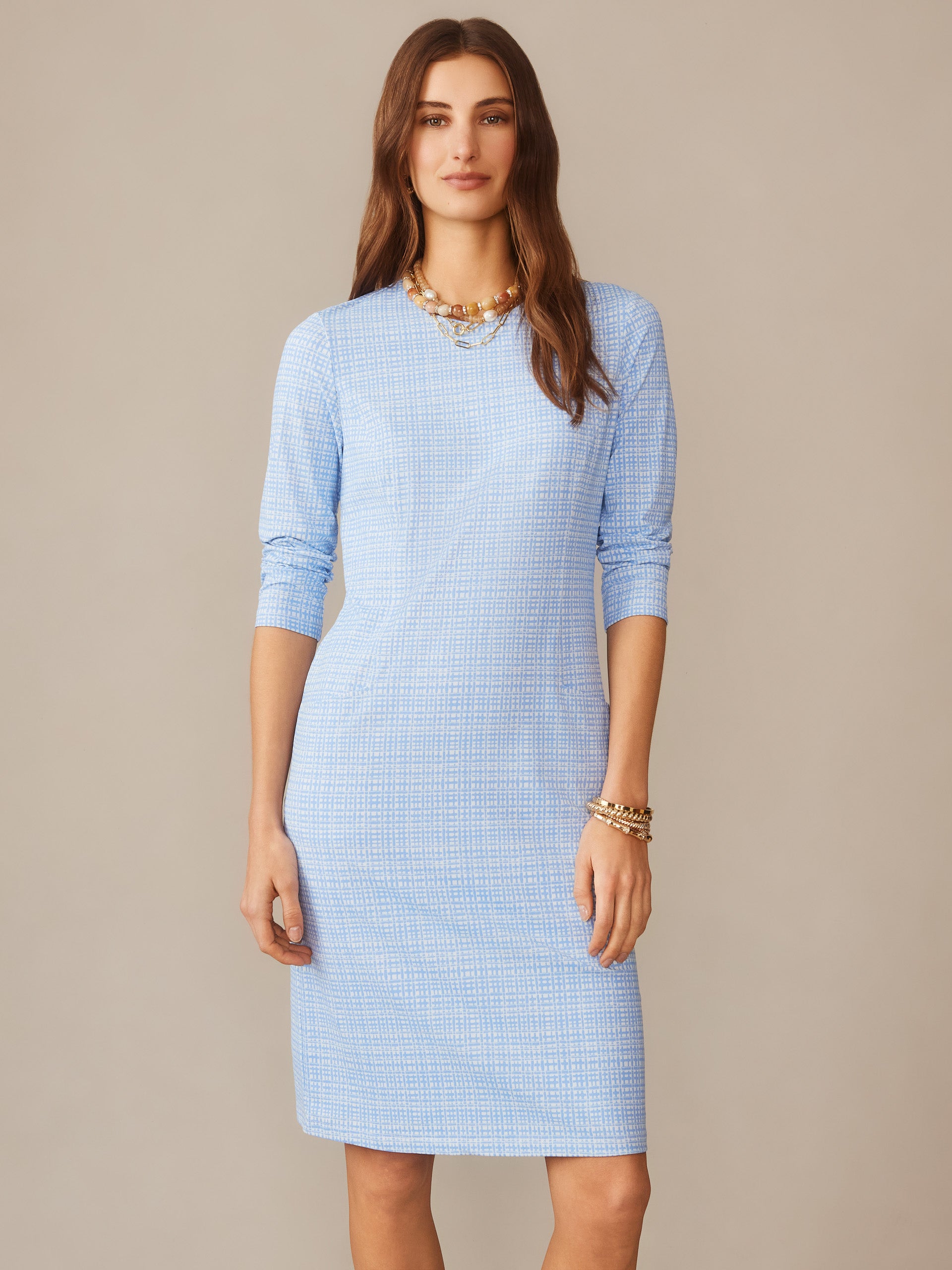 Woman wearing a light blue patterned Catalyst Dress in Pocket Square by J.McLaughlin, styled with layered gold necklaces and bracelets, posing against a neutral background.