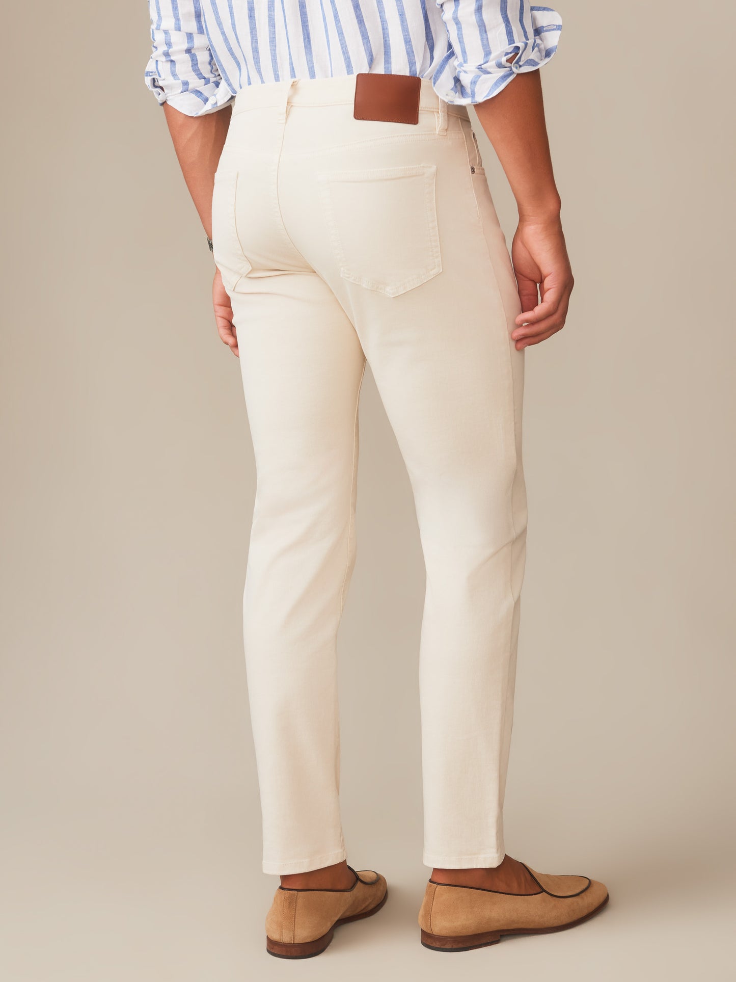 A rear view of a person wearing cream-colored Mercer Pants by J.McLaughlin, paired with casual loafers and a striped shirt, showcasing effortlessly stylish, enduring designs in innovative fabrics. A destination for defining style. Life, Well-Lived.