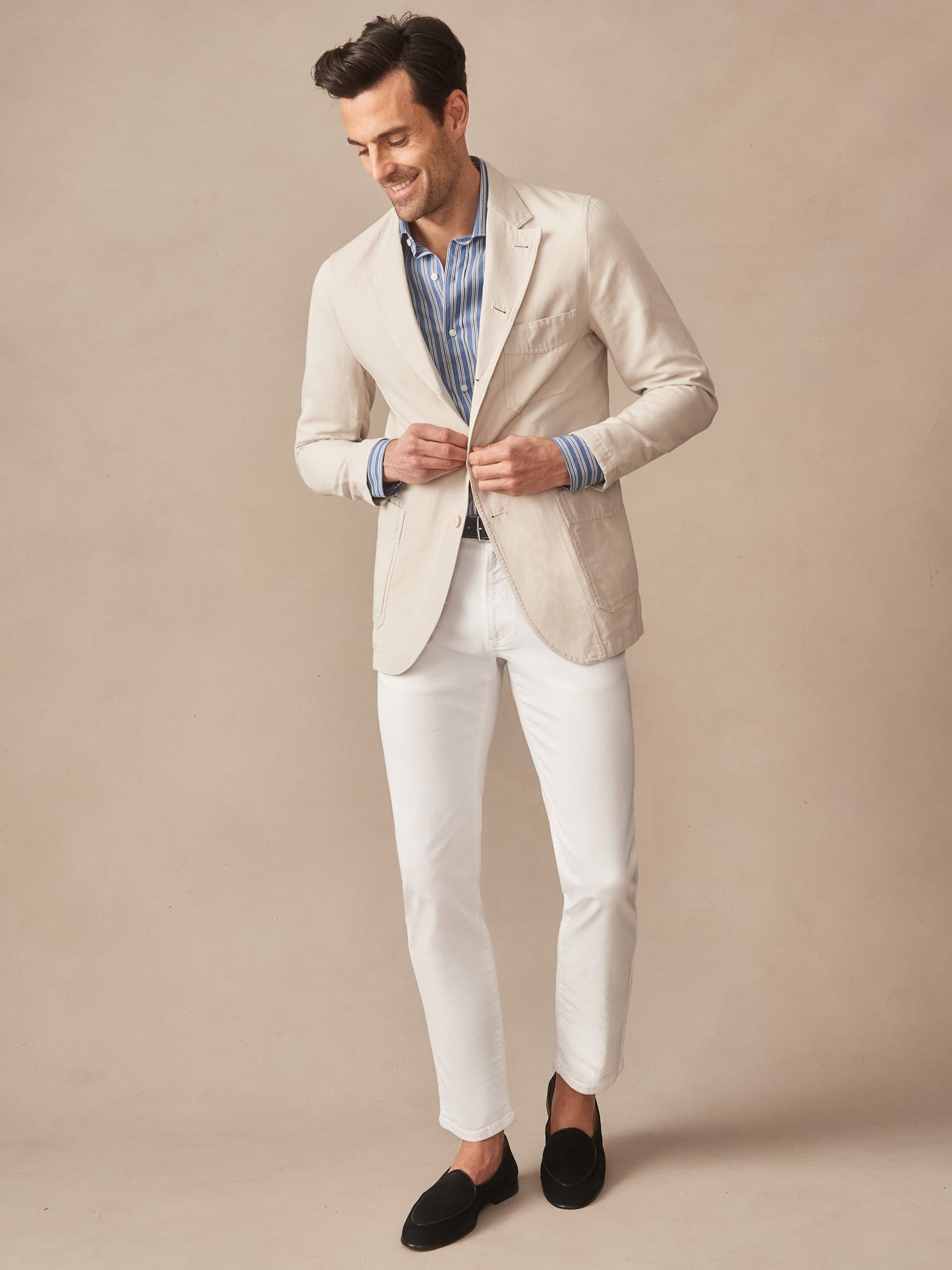 Man wearing a beige Charlestown Jacket by J.McLaughlin over a blue striped shirt with white pants and black loafers, showcasing effortlessly stylish, enduring designs with innovative fabrics.