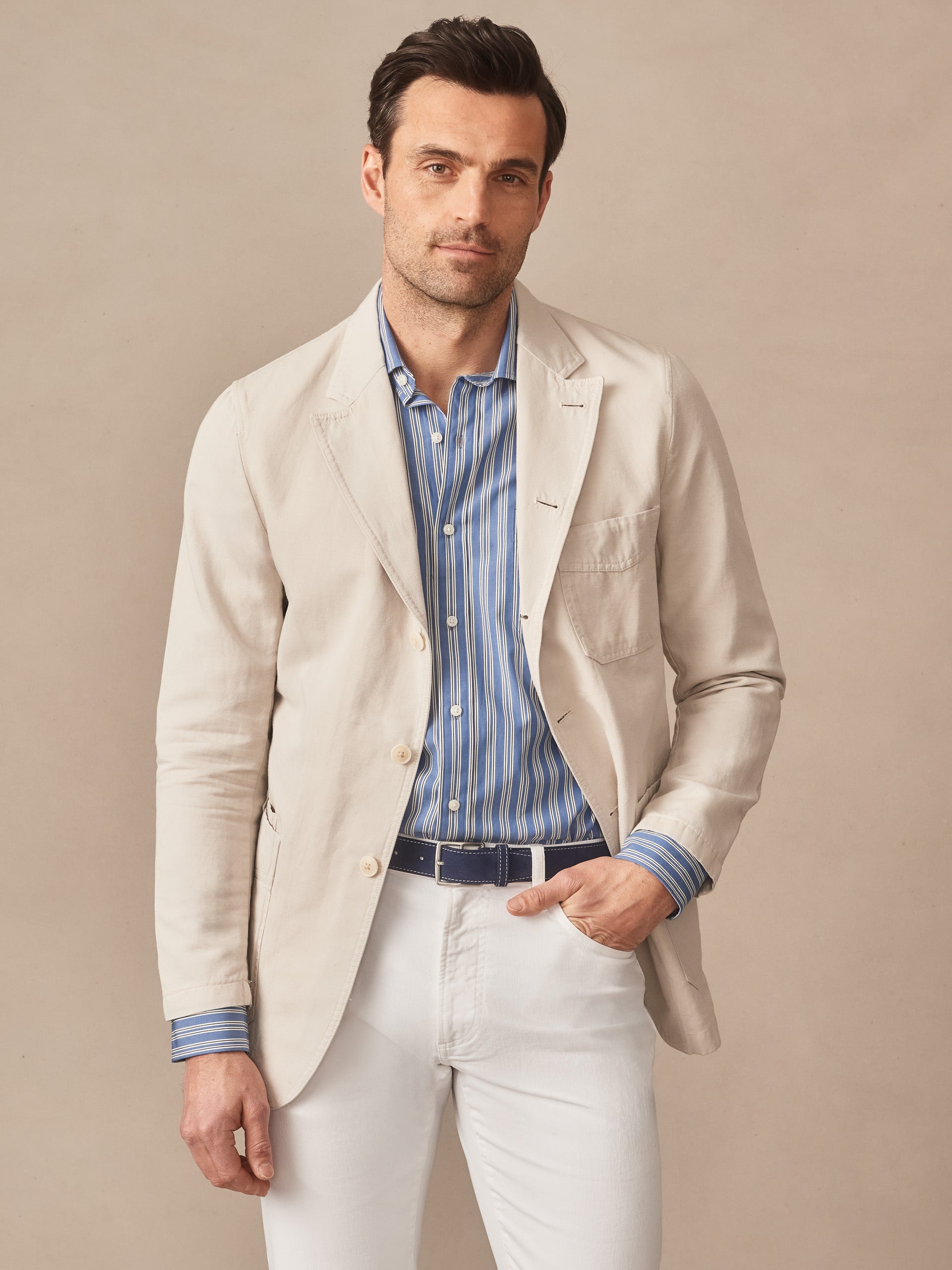 Man wearing a J.McLaughlin Charlestown Jacket in light beige over a blue striped shirt and white pants, showcasing effortlessly stylish, enduring designs and innovative fabrics in timeless fashion.
