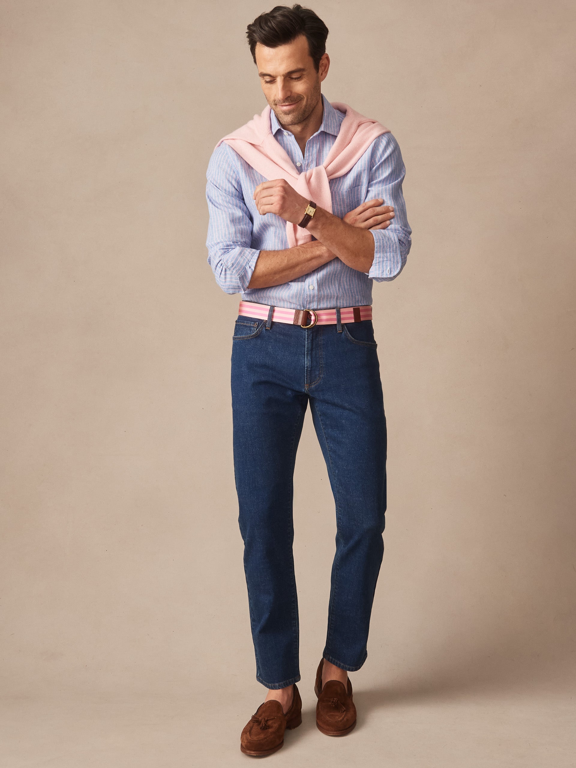 Man wearing Mercer Jeans by J.McLaughlin in dark denim paired with a striped button-down shirt, pink sweater draped over shoulders, and brown tassel loafers, showcasing effortlessly stylish, enduring designs and innovative fabrics. A destination for defining style. Life, Well-Lived.