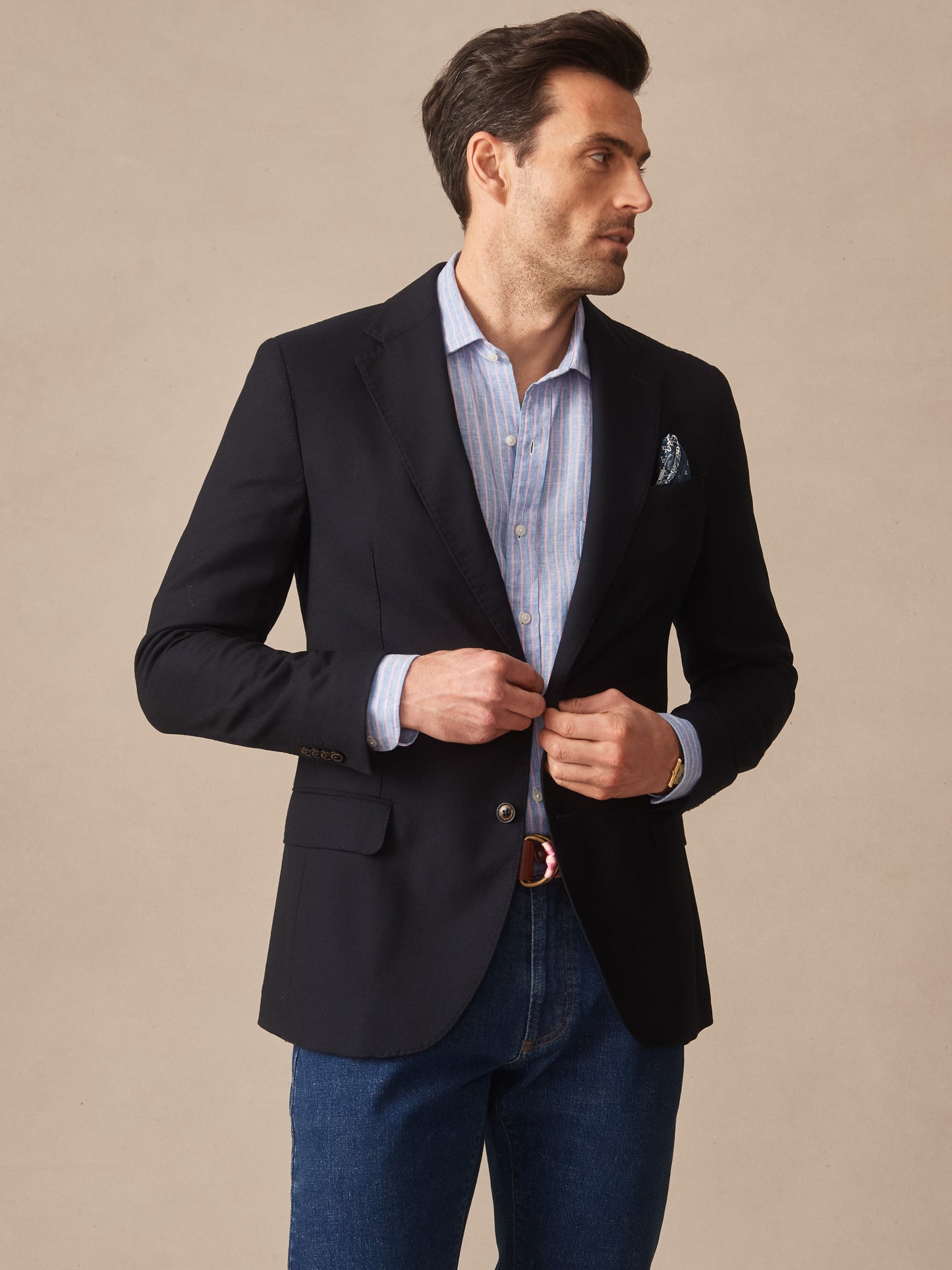 Man wearing a dark Suffolk Sport Coat by J.McLaughlin over a light blue striped shirt and denim jeans, showcasing effortlessly stylish, enduring designs and innovative fabrics.