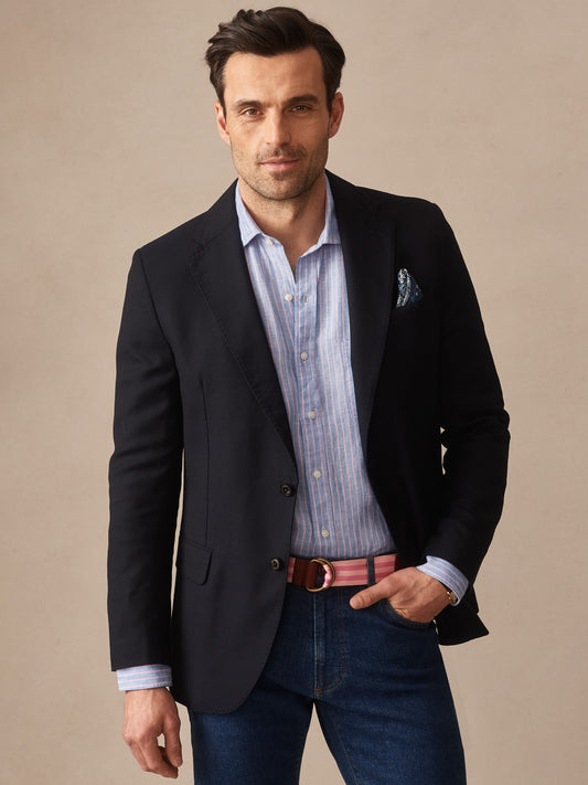 Man wearing a navy Suffolk Sport Coat by J.McLaughlin over a light blue striped shirt with dark denim jeans and a pink belt, showcasing effortlessly stylish, enduring designs and innovative fabrics.