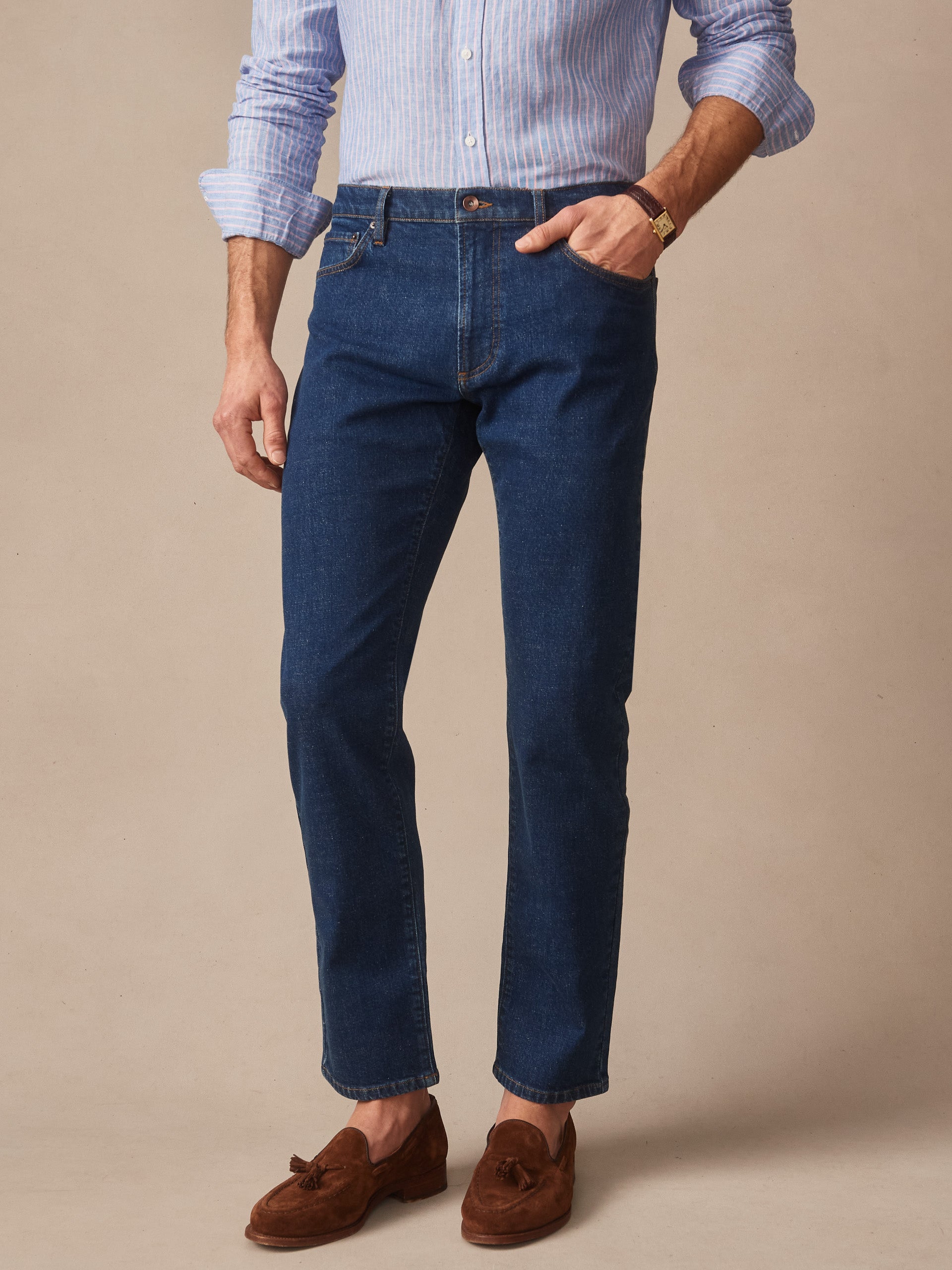 Man wearing dark denim Mercer Jeans by J.McLaughlin paired with a light blue striped shirt and brown suede loafers, showcasing effortlessly stylish, enduring designs and innovative fabrics.