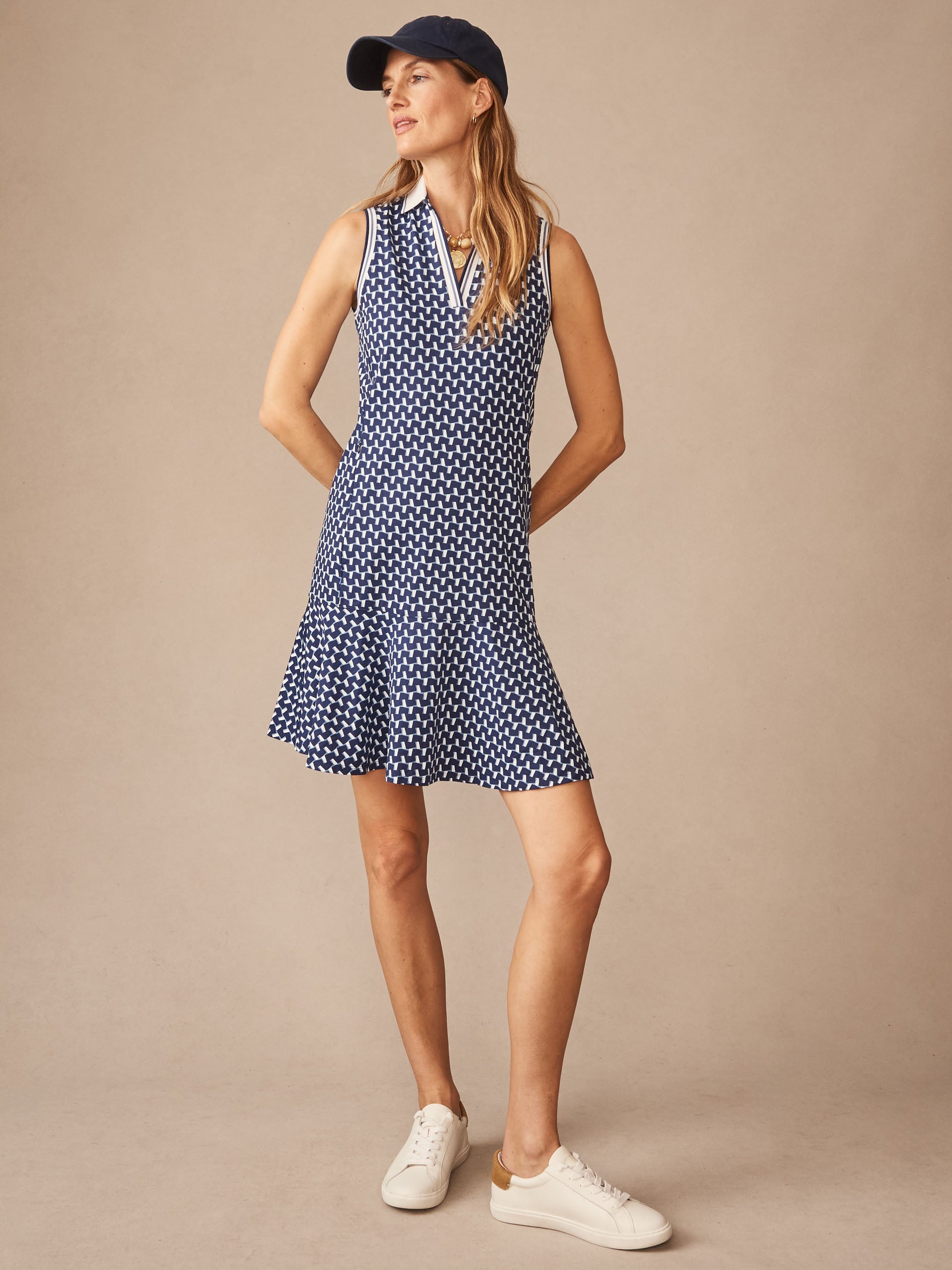 Woman wearing the Merlin Dress in Duncan by J.McLaughlin, a sleeveless navy and light blue patterned dress paired with white sneakers and a navy cap, embodying effortlessly stylish, enduring designs with a legacy of timeless fashion. A destination for defining style. Life, Well-Lived.