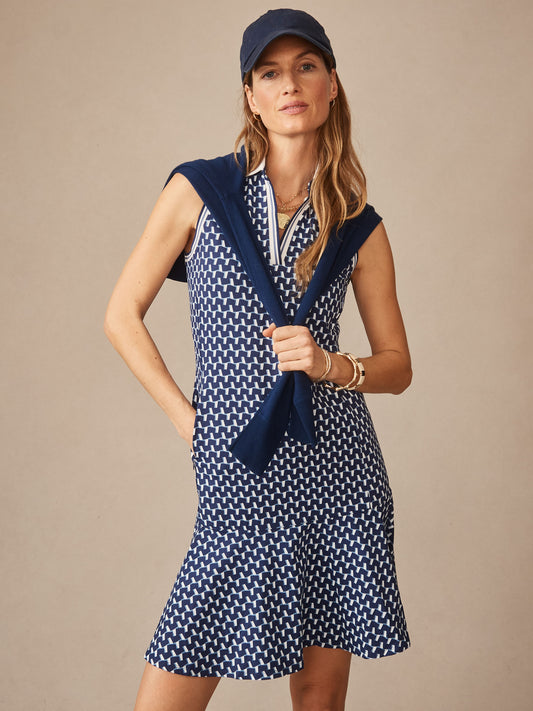 Woman wearing the Merlin Dress in Duncan by J.McLaughlin, featuring a navy and light blue geometric pattern with a sleeveless design, styled with a navy sweater draped over her shoulders and matching cap, showcasing effortlessly stylish, enduring designs and innovative fabrics, a destination for defining style. Life, Well-Lived.