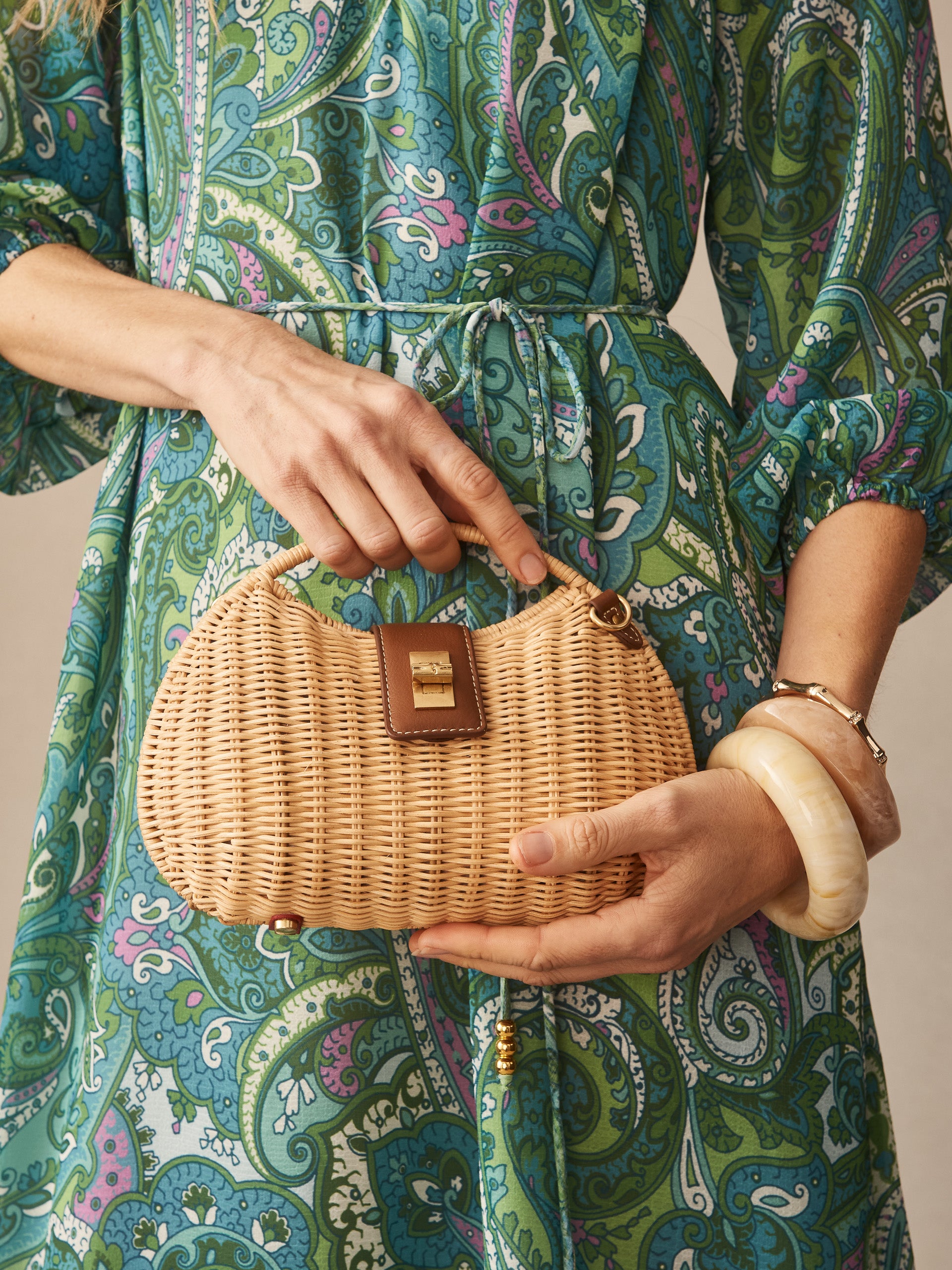 A woman wearing a paisley print dress holds an Iris Wicker Handbag by J.McLaughlin, showcasing effortlessly stylish, enduring designs and a legacy of timeless fashion.
