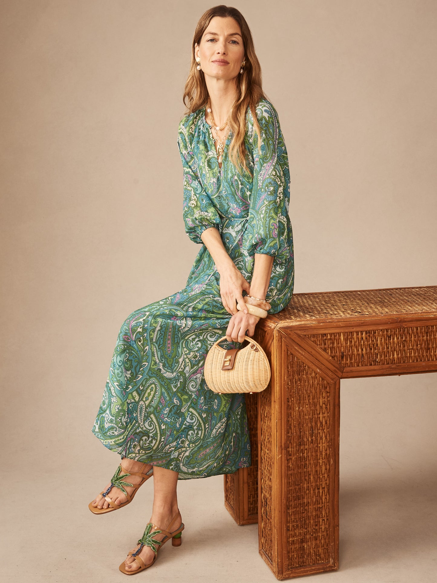 Woman wearing the Mia Dress in Paisley Reverie by J.McLaughlin, featuring a flowing paisley print in blue and green, sitting on a woven wooden bench holding a small woven handbag, effortlessly stylish and enduring designs embodying the legacy of timeless fashion at a destination for defining style. Life, Well-Lived.