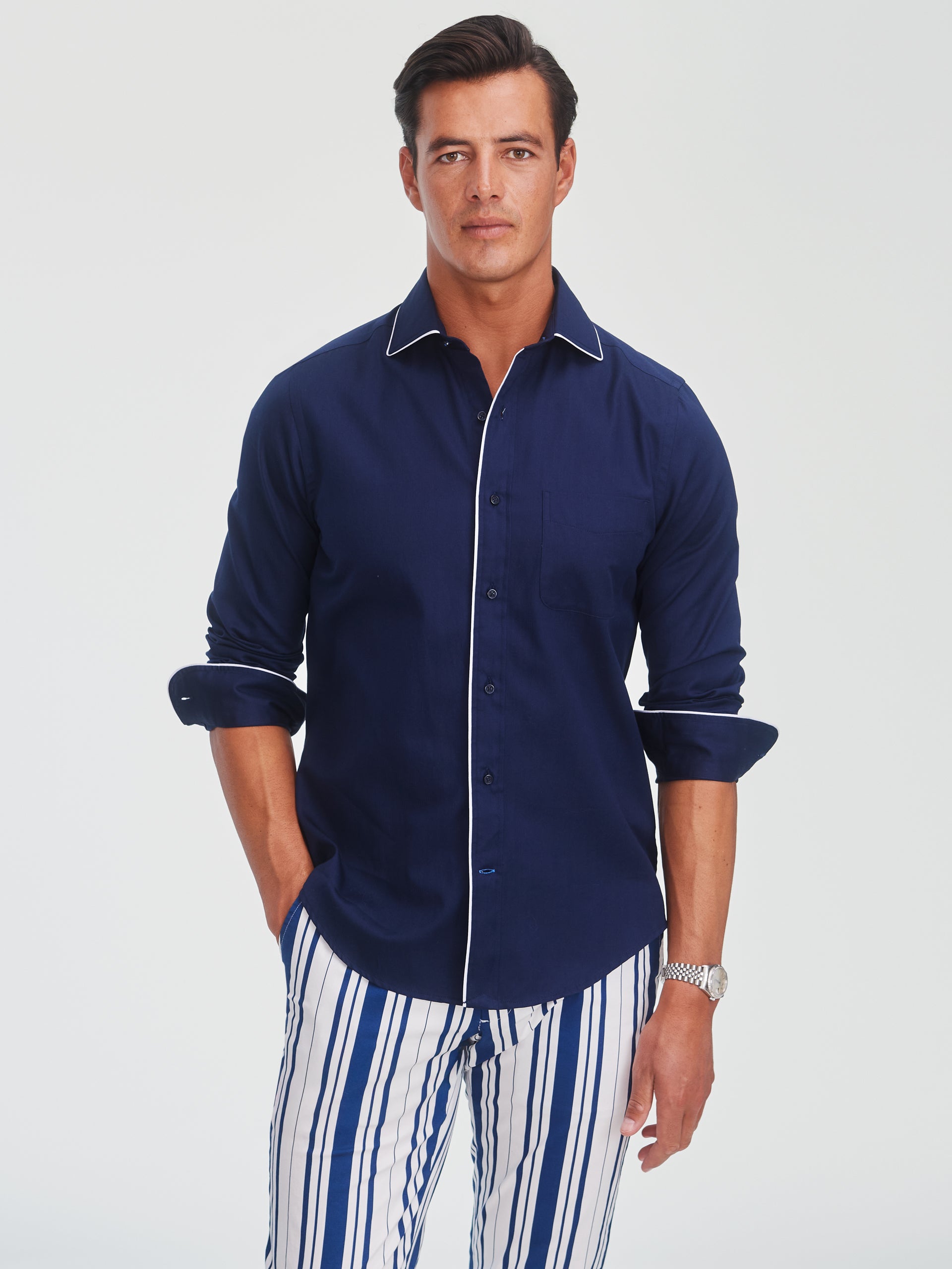 A man wearing the Anderson Cotton Shirt by J.McLaughlin in navy with white piping, paired with striped pants, showcasing effortlessly stylish, enduring designs, innovative fabrics, and a legacy of timeless fashion. A destination for defining style. Life, Well-Lived.