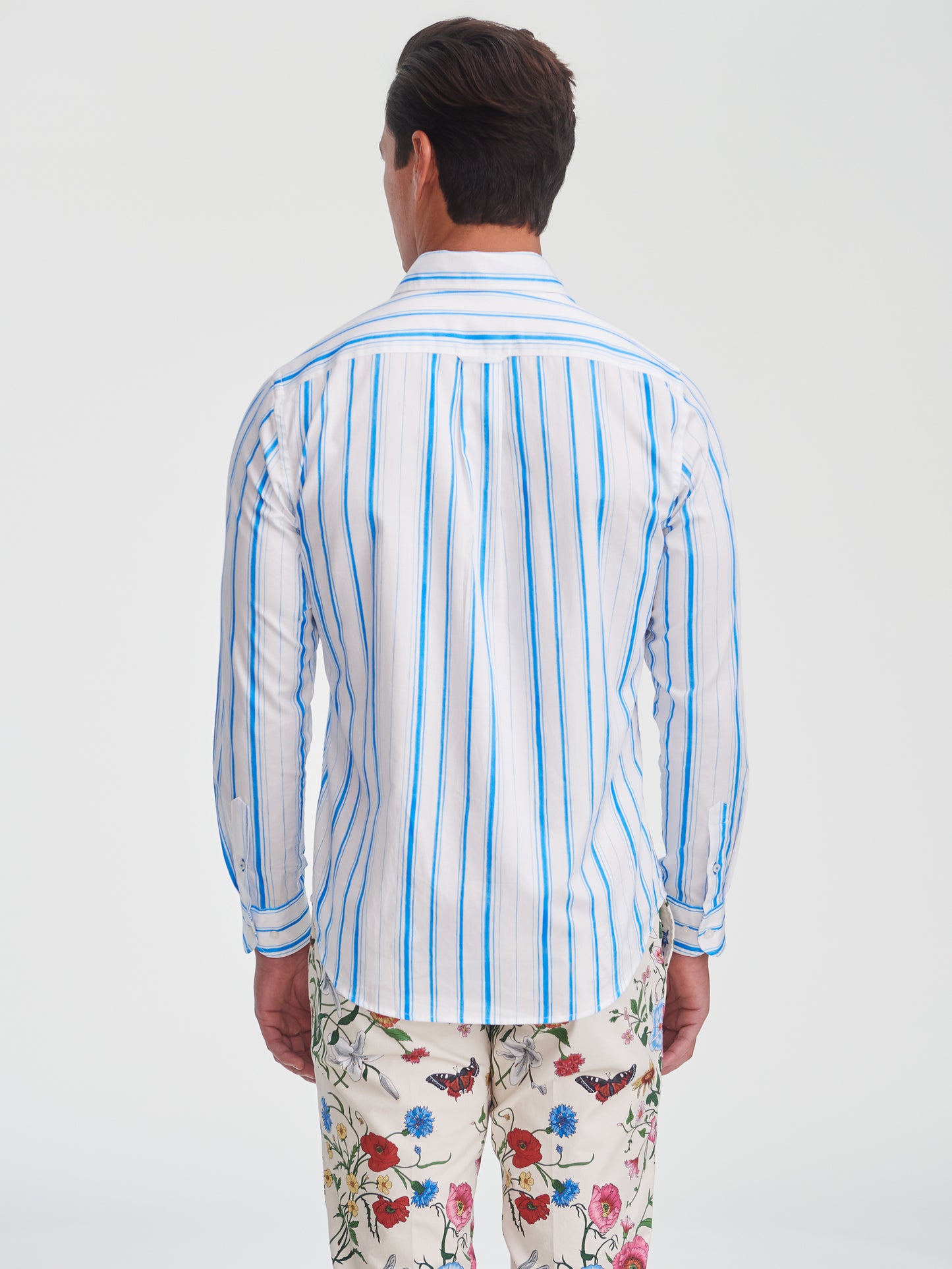 Man wearing the Gramercy Cotton Shirt in Coastal Waterline by J.McLaughlin, featuring blue and white stripes, paired with floral print pants, showcasing effortlessly stylish, enduring designs and innovative fabrics. A destination for defining style. Life, Well-Lived.