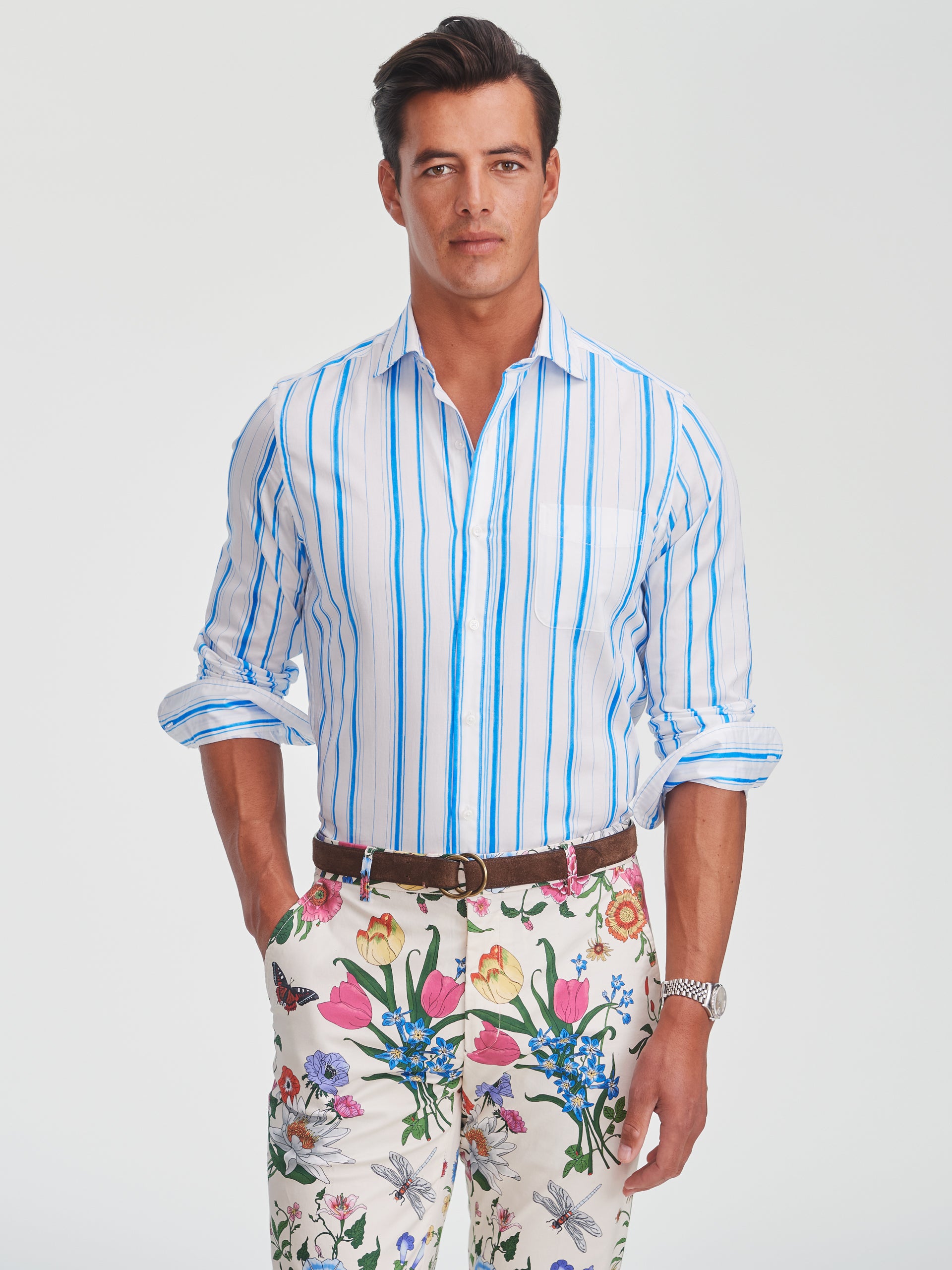 A man wearing the Gramercy Cotton Shirt in Coastal Waterline by J.McLaughlin, featuring blue vertical stripes on white cotton, paired with floral patterned pants, showcasing effortlessly stylish, enduring designs and innovative fabrics. A destination for defining style. Life, Well-Lived.