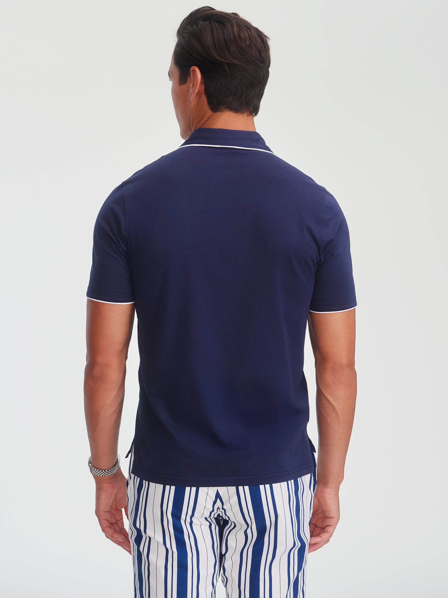 Man wearing a navy Ryan Polo by J.McLaughlin with contrasting white trim on the sleeves and collar, paired with striped pants, showcasing effortlessly stylish, enduring designs and innovative fabrics.