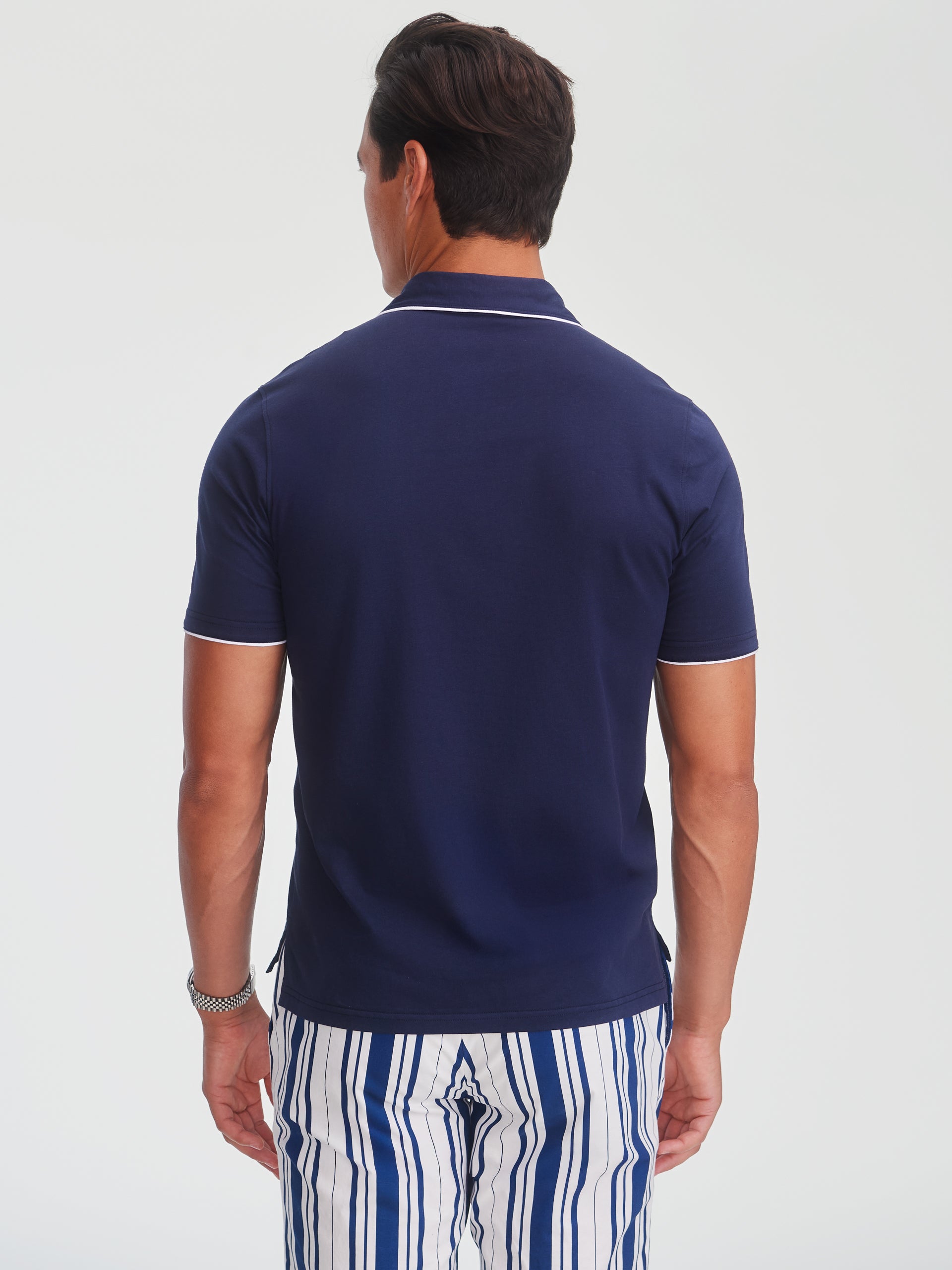 Man wearing a navy Ryan Polo by J.McLaughlin with contrasting white trim on the sleeves and collar, paired with striped pants, showcasing effortlessly stylish, enduring designs and innovative fabrics.