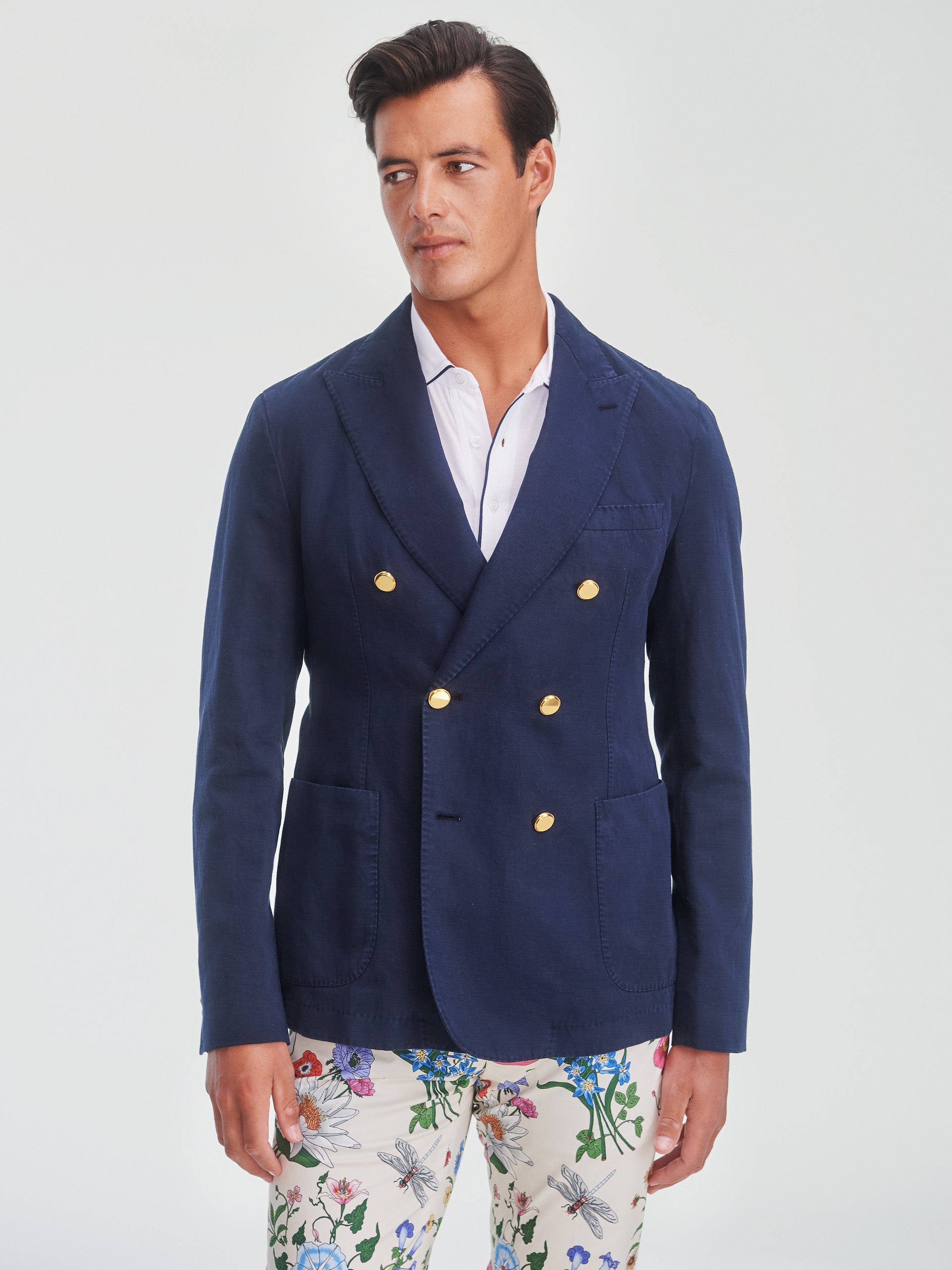 A man wearing a navy double-breasted Mark Sport Coat with gold buttons and a white shirt, paired with floral patterned pants, showcasing J.McLaughlin’s effortlessly stylish, enduring designs and innovative fabrics. A destination for defining style. Life, Well-Lived.