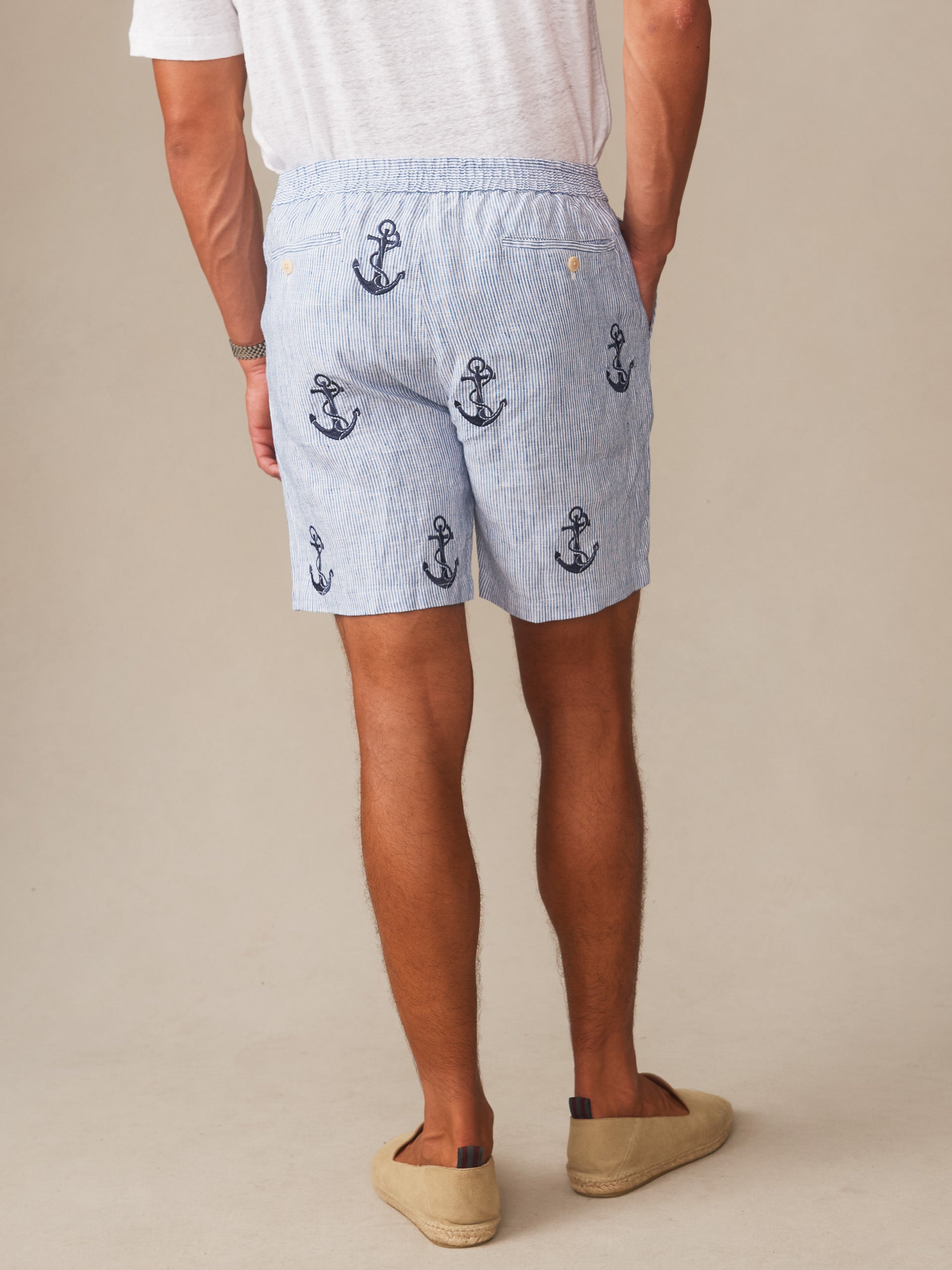 A rear view of a man wearing J.McLaughlin Newport Linen Shorts in Anchor adorned with navy anchor motifs, paired with casual slip-on shoes, showcasing effortlessly stylish, enduring designs crafted from innovative fabrics. A destination for defining style. Life, Well-Lived.