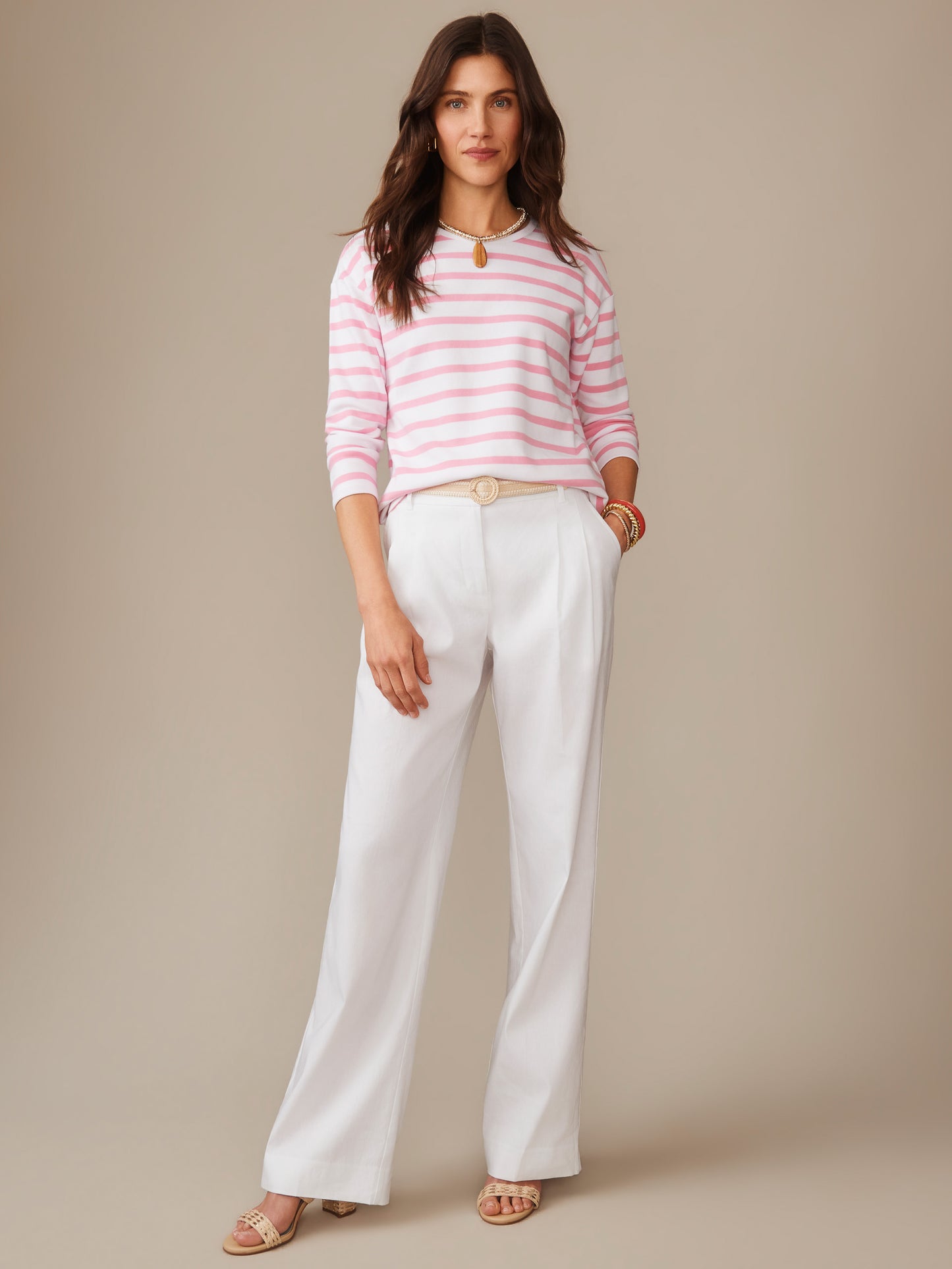 A woman models the Nico Tee in Stripe by J.McLaughlin, featuring pink and white stripes paired with white pants, showcasing effortlessly stylish, enduring designs in innovative fabrics.