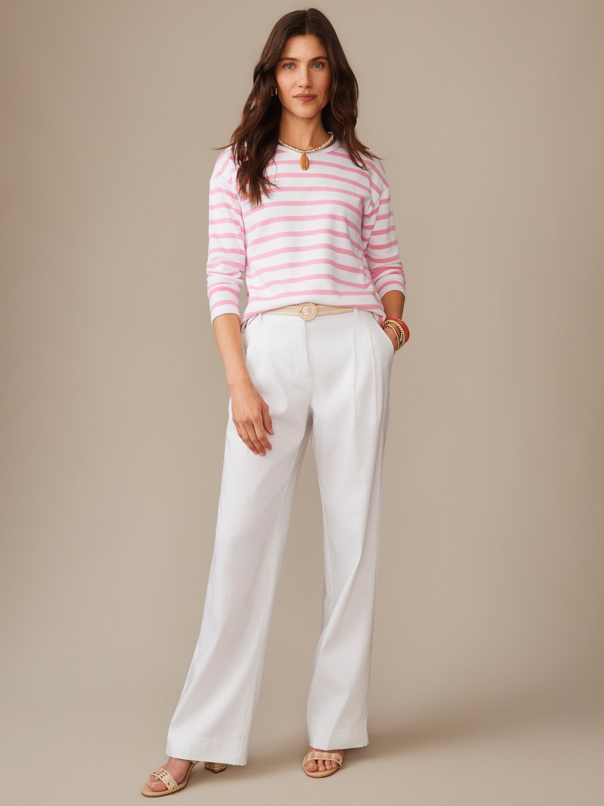 A woman models the Nico Tee in Stripe by J.McLaughlin, featuring pink and white stripes paired with white pants, showcasing effortlessly stylish, enduring designs in innovative fabrics.